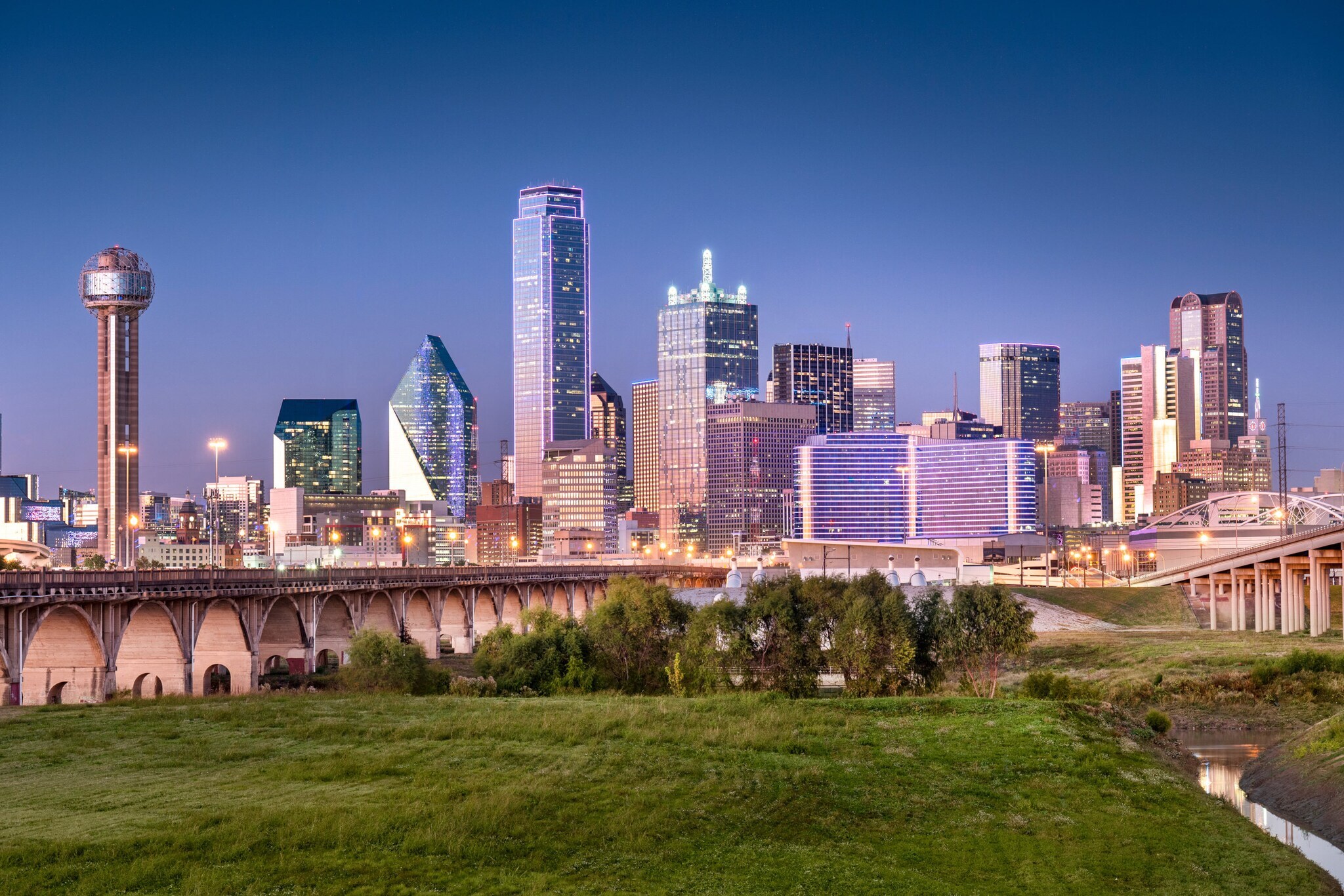 Skyline von Dallas mit Wolkenkratzern hinter einer Grünfläche mit Brückenbauten bei Abenddämmerung. Skyline von Dallas mit Wolkenkratzern hinter einer Grünfläche mit Brückenbauten bei Abenddämmerung.