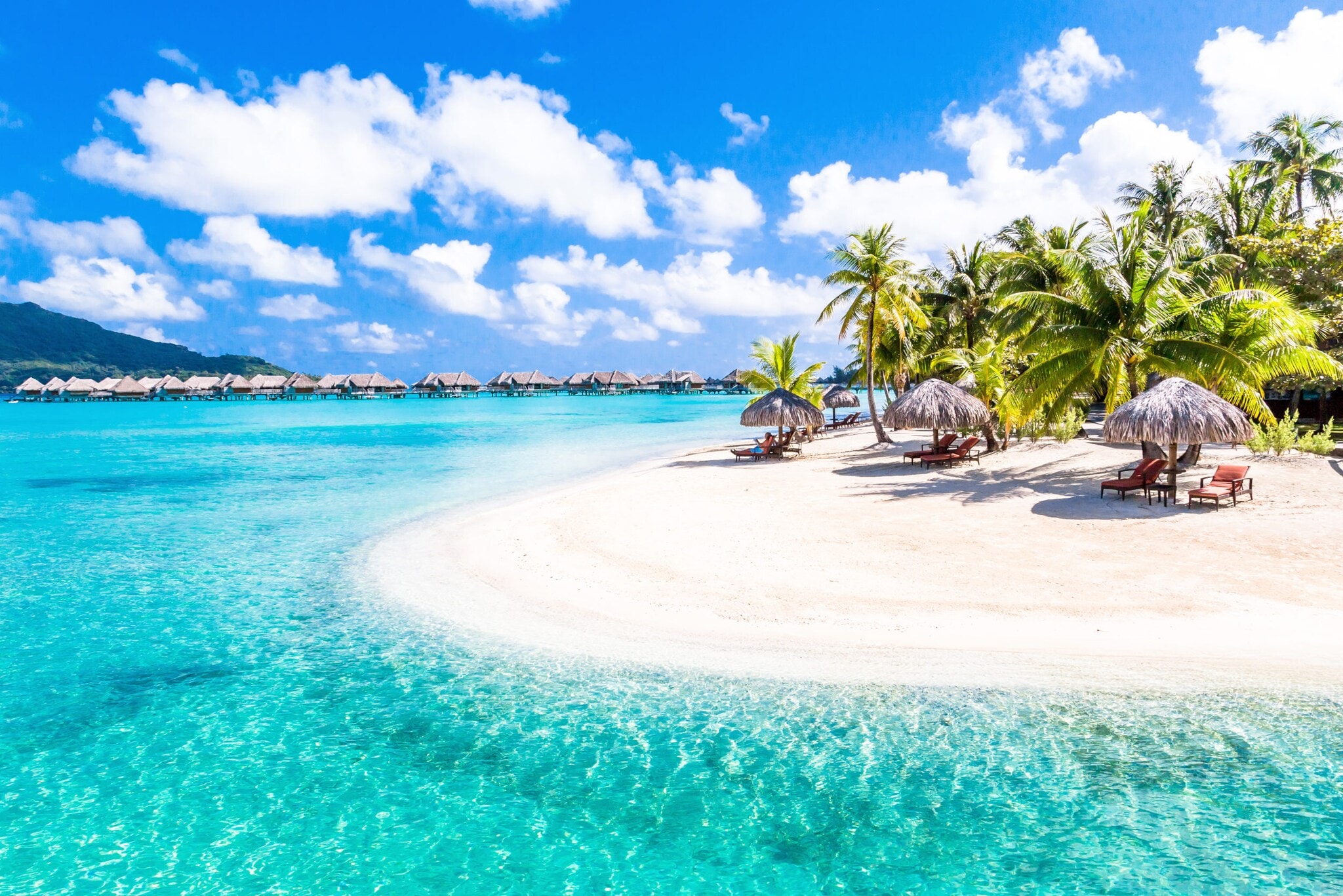 Strandliegen mit Strohschirmen auf einem palmengesäumten Strand mit weißem Sand an türkisblauem Wasser, im Hintergrund Hotelresort mit Überwasser-Bungalows. Strandliegen mit Strohschirmen auf einem palmengesäumten Strand mit weißem Sand an türkisblauem Wasser, im Hintergrund Hotelresort mit Überwasser-Bungalows.