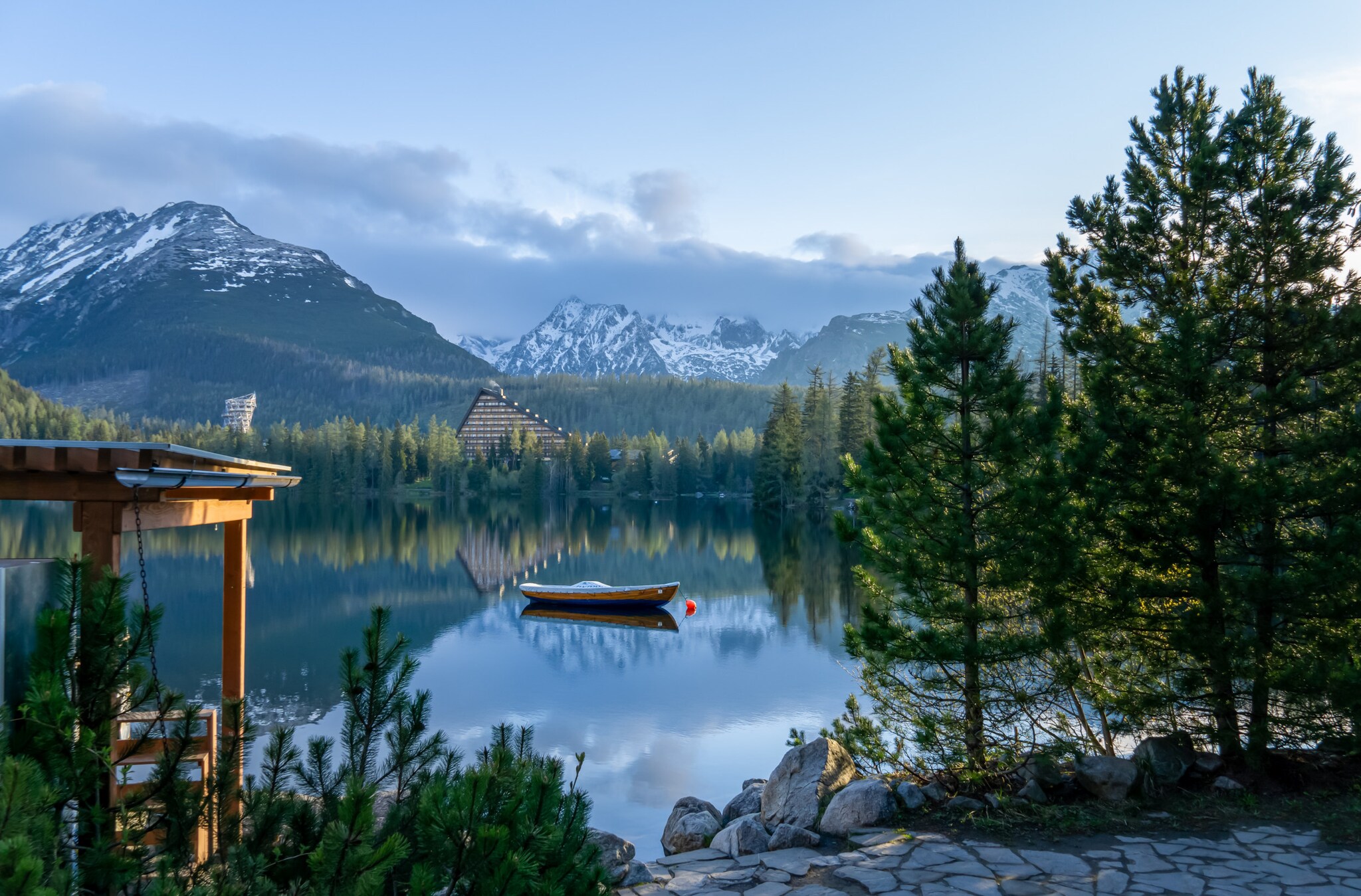 Ein idyllischer Bergsee mit Boot, umgeben von Nadelwald in der Abenddämmerung, im Hintergrund schneebedecktes Gipfelpanorama.