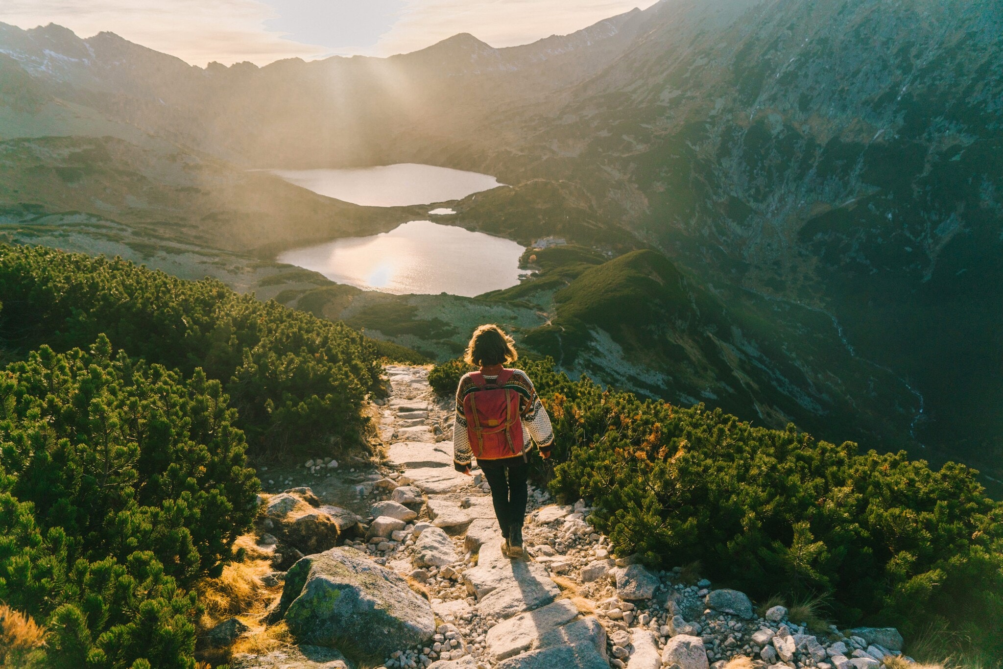 Rückansicht einer Frau im Pullover mit Rucksack auf einem Wanderweg im grünen Gebirge vor zwei Bergseen im Gegenlicht. Rückansicht einer Frau im Pullover mit Rucksack auf einem Wanderweg im grünen Gebirge vor zwei Bergseen im Gegenlicht.