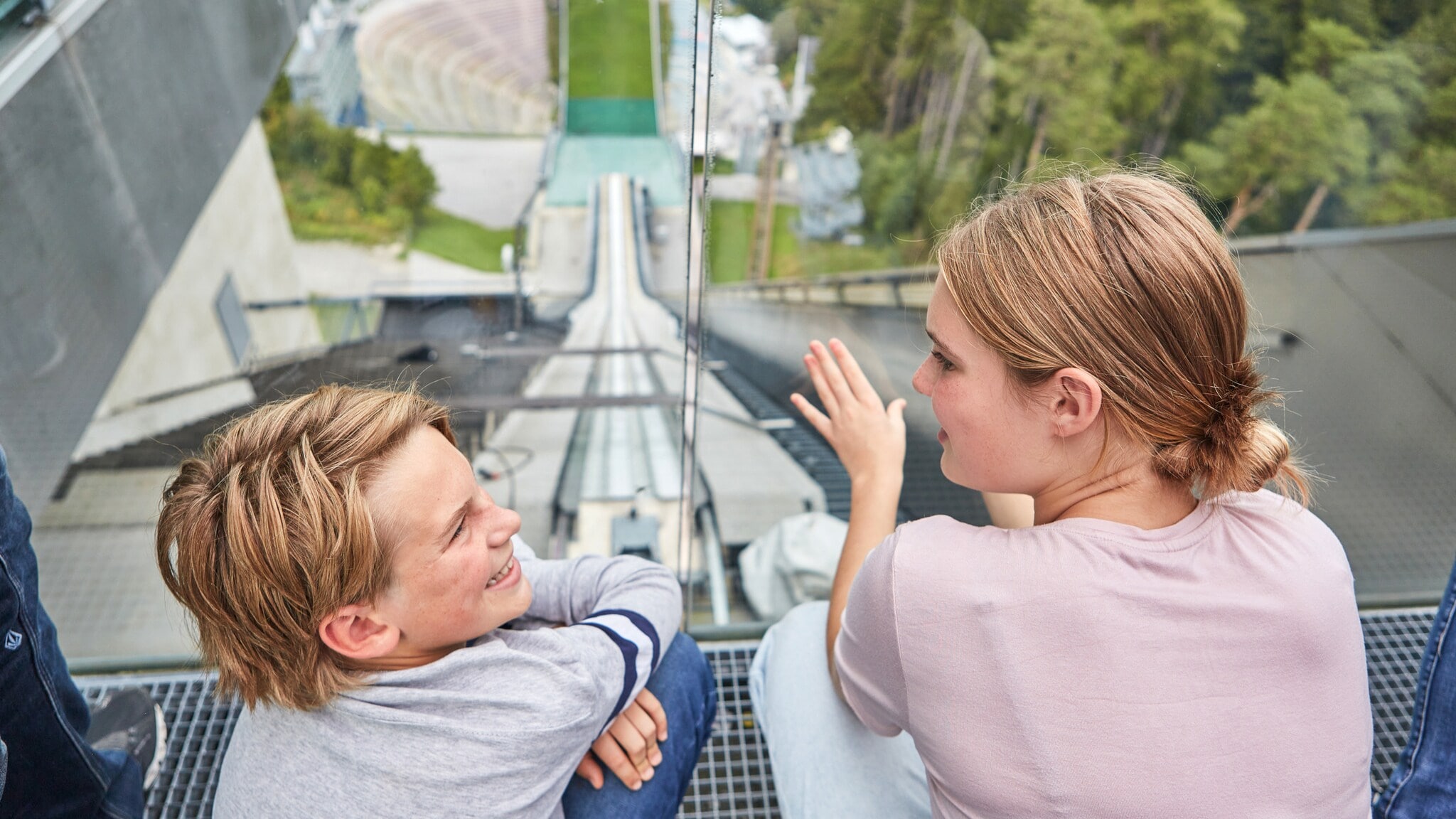 Zwei Kinder im Profil an einer Glasscheibe auf einer Aussichtsplattform einer Sprungschanze im Sommer. Zwei Kinder im Profil an einer Glasscheibe auf einer Aussichtsplattform einer Sprungschanze im Sommer.