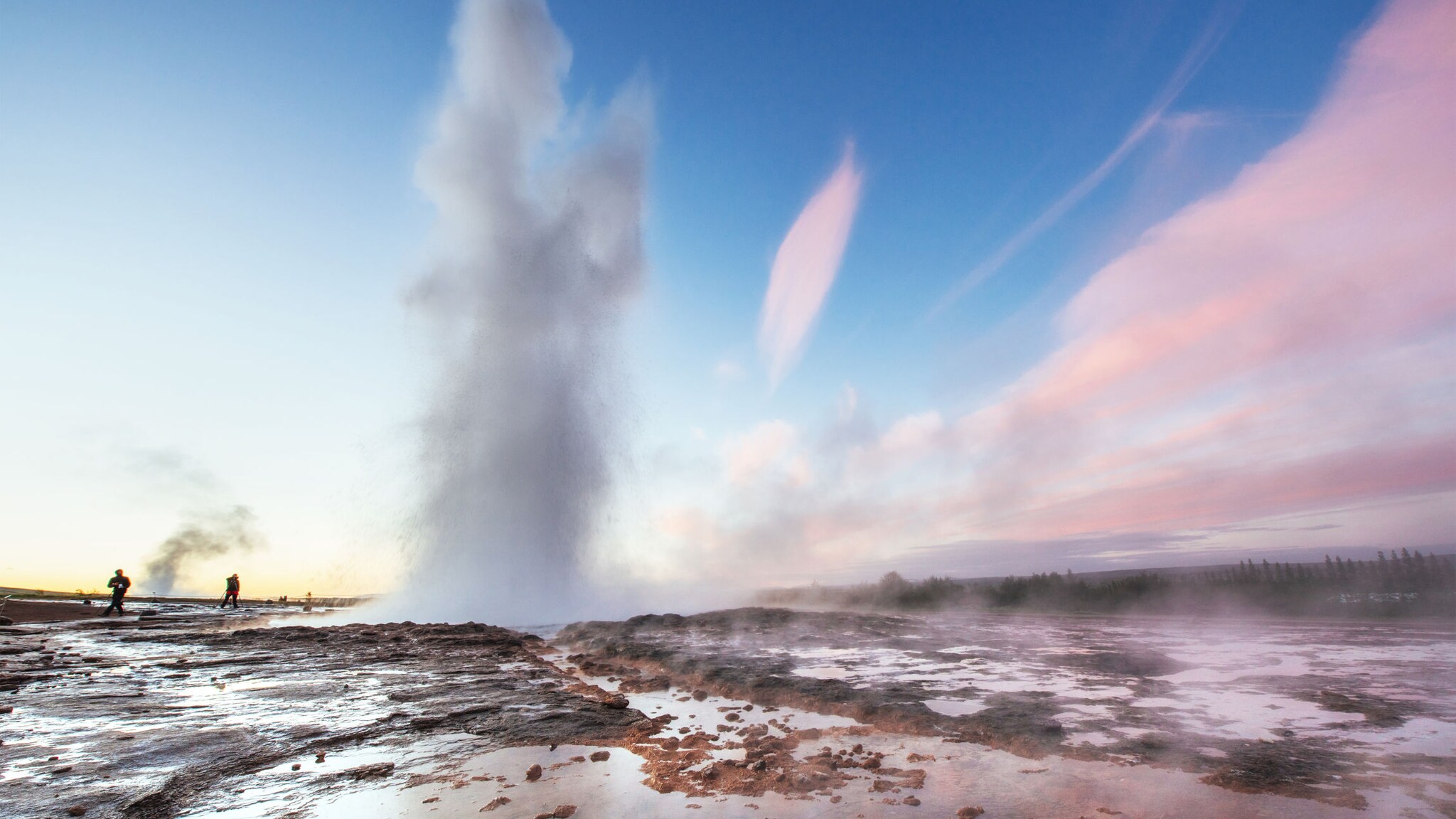 Ein sprudelnder Geysir auf einem Felsplateau unter blauem Himmel, im Hintergrund vereinzelte Personen.