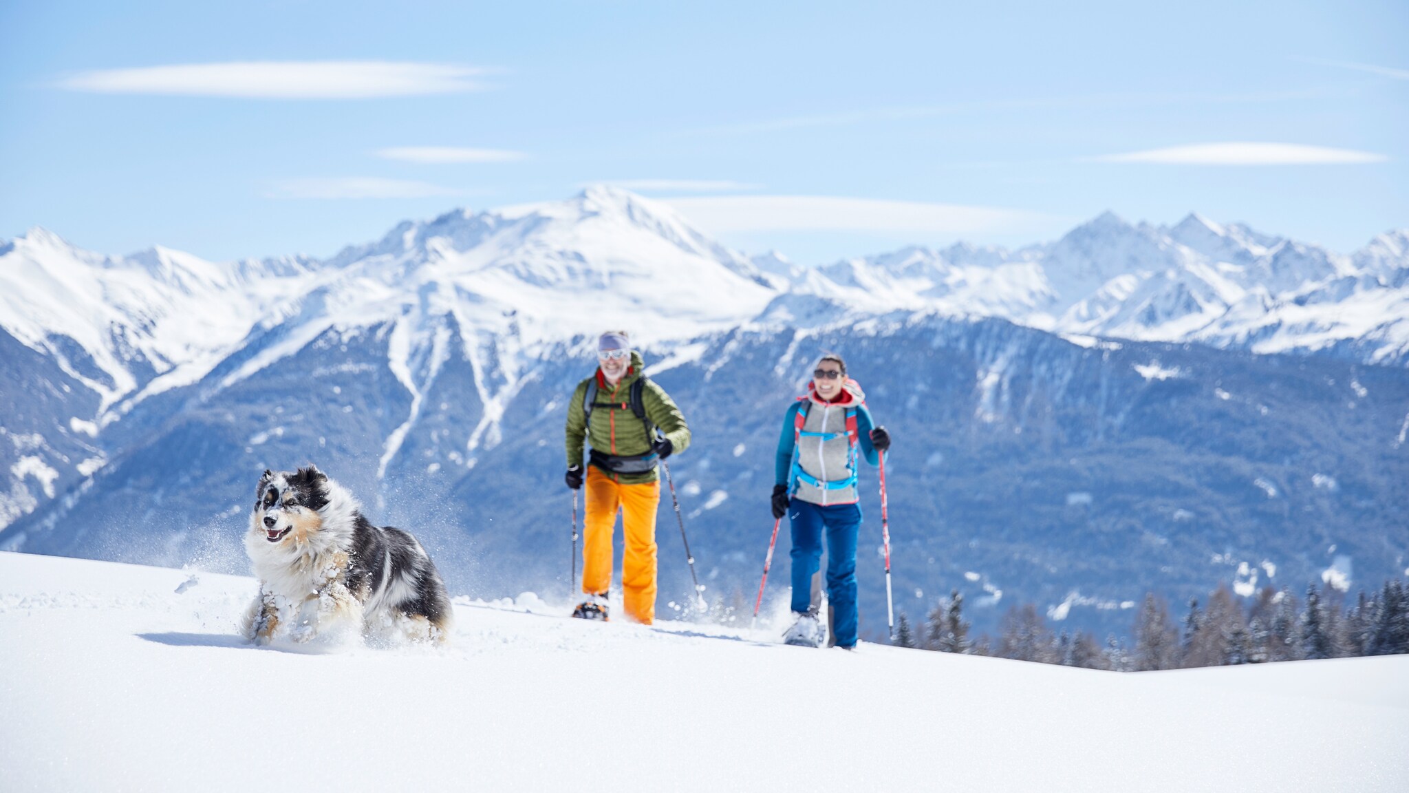 Ein Paar beim Schneeschuhwandern mit einem Hund vor schneebedecktem Gipfelpanorama.