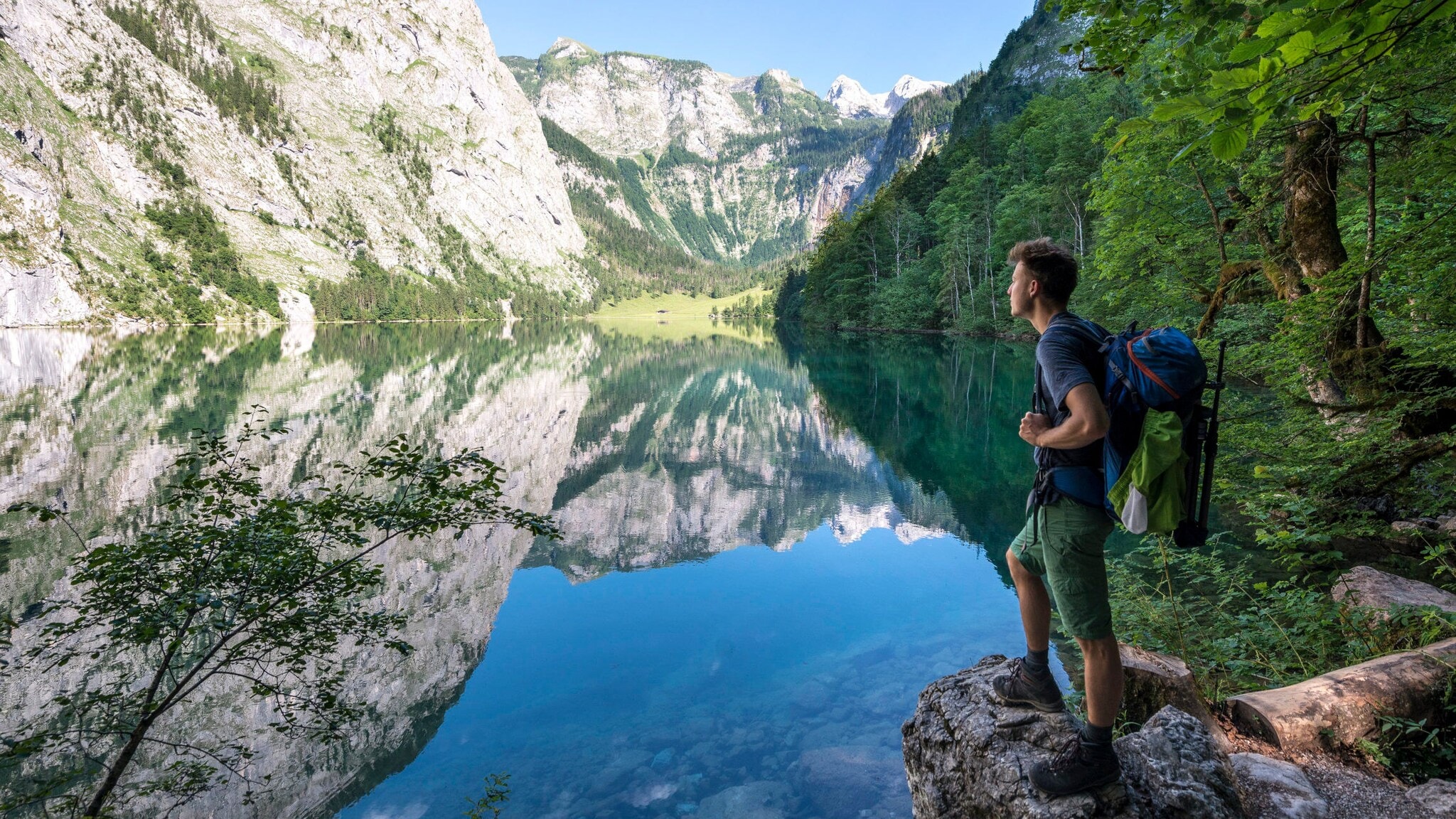 Eine Person mit Wanderrucksack steht auf einem Stein vor dem Königssee mit Blick auf die Berge. Eine Person mit Wanderrucksack steht auf einem Stein vor dem Königssee mit Blick auf die Berge.
