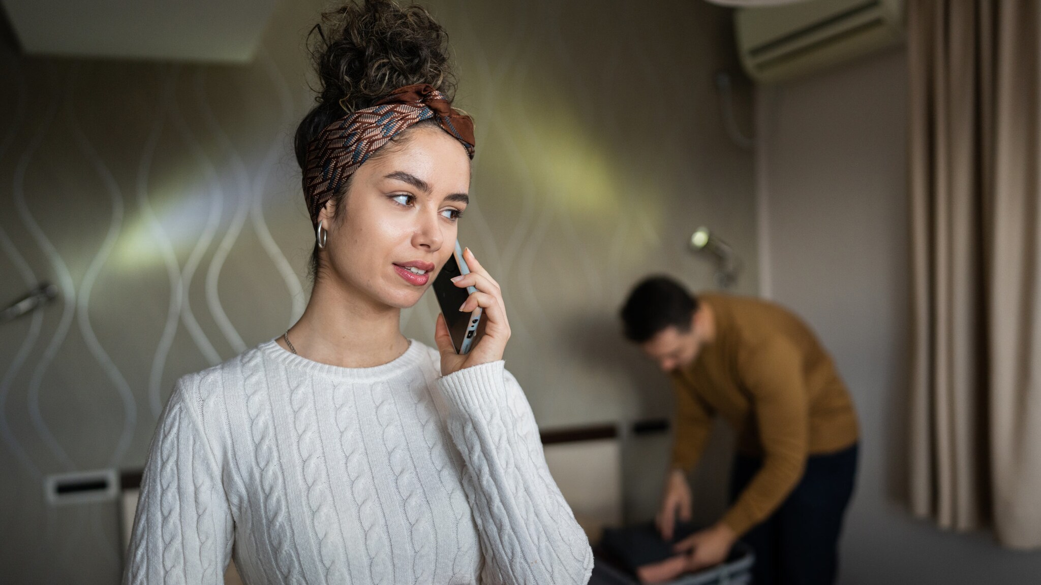 Eine Frau, die ein Smartphone an ihr Ohr hält, im Hintergrund steht ein Mann über einen Koffer gebeugt.