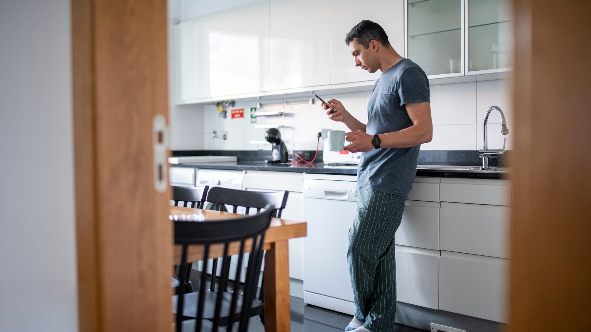 Ein Mann steht in einer Küche und hält in einer Hand eine Tasse und in der anderen ein Smartphone. Ein Mann steht in einer Küche und hält in einer Hand eine Tasse und in der anderen ein Smartphone.