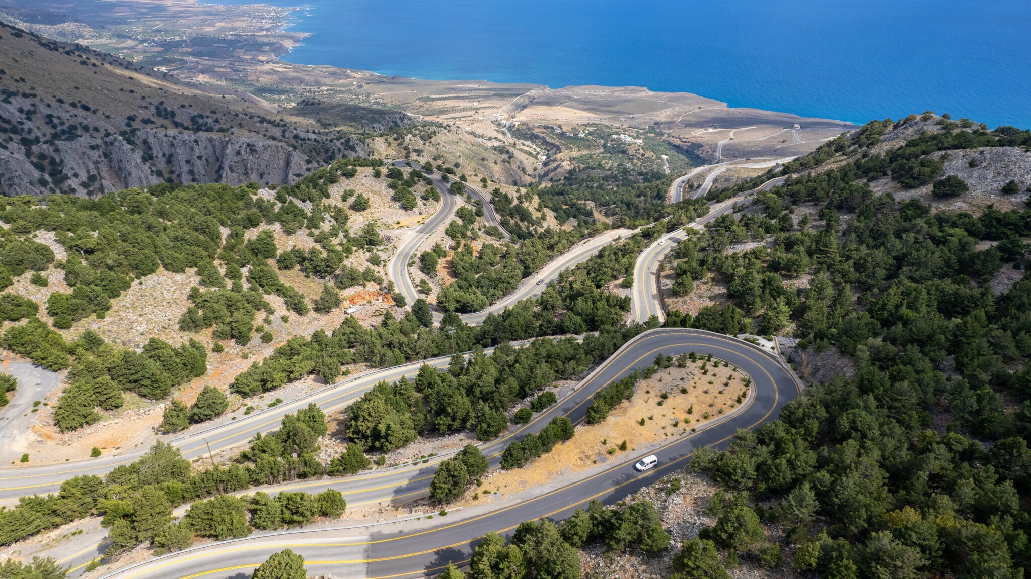 Ein Auto auf einer Serpentinenstraße in einer mediterranen Berglandschaft an einer Meeresküste.