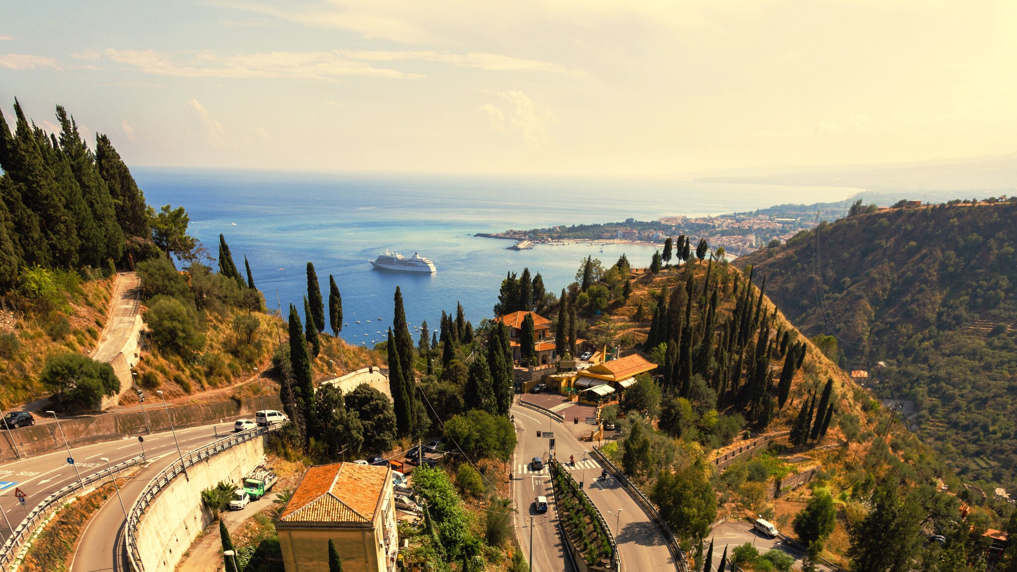 Serpentinenstraße in mediterraner Berglandschaft mit Zypressen mit Blick auf eine Meeresbucht mit Kreuzfahrtschiff.