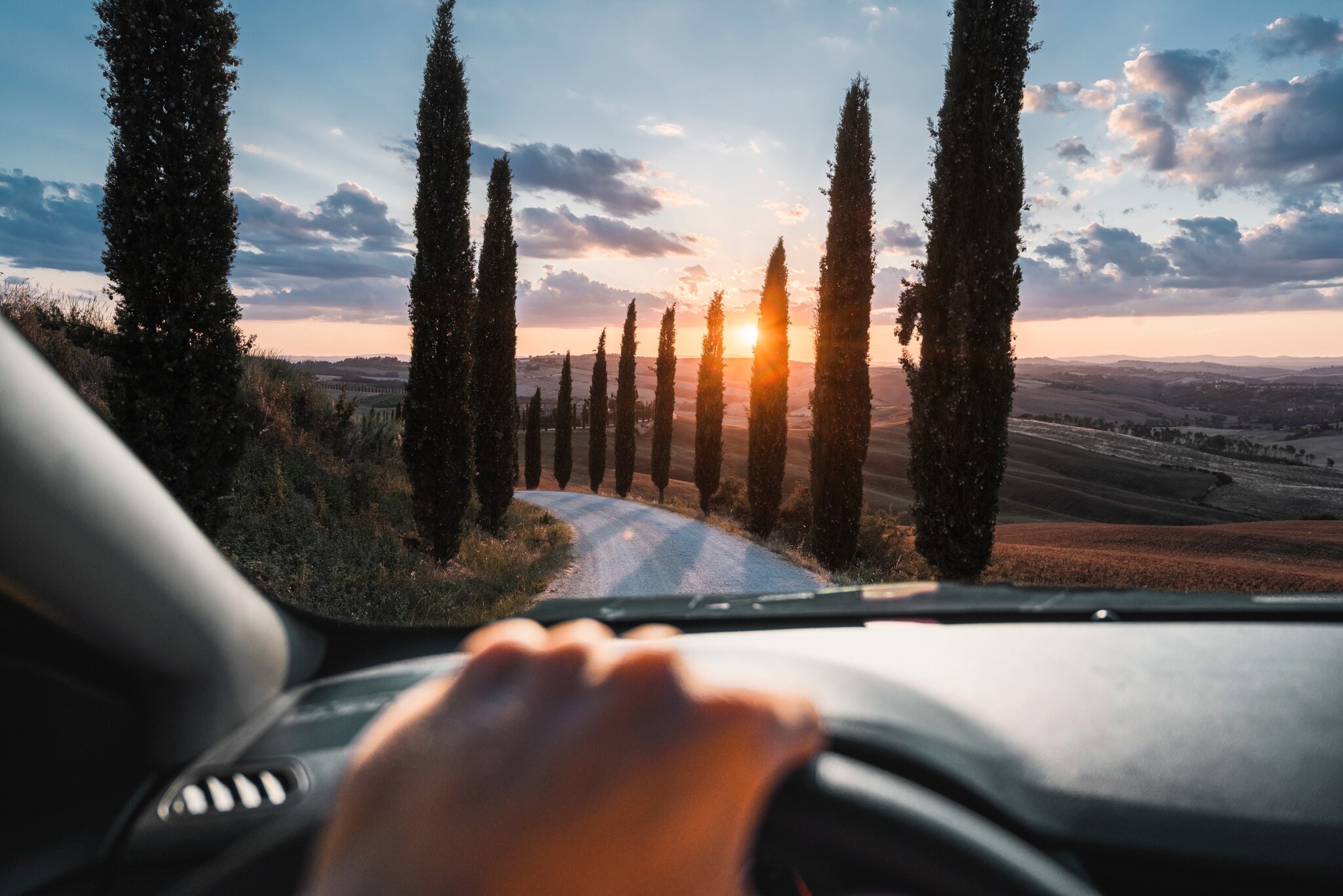 Blick auf eine mit Zypressen gesäumte Landstraße in Hügellandschaft bei Sonnenuntergang durch die Frontscheibe eines Autos, im Vordergrund eine Hand am Lenkrad. Blick auf eine mit Zypressen gesäumte Landstraße in Hügellandschaft bei Sonnenuntergang durch die Frontscheibe eines Autos, im Vordergrund eine Hand am Lenkrad.