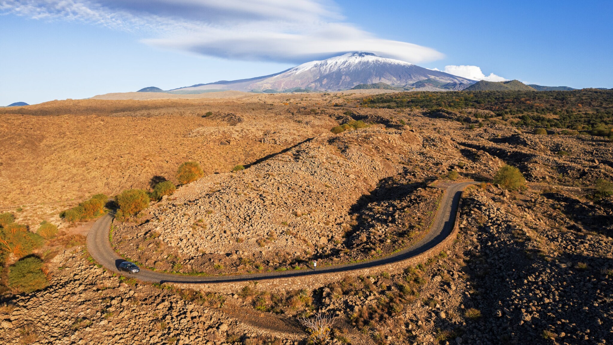 Ein Auto fährt auf einer kurvigen Landstraße in karger Landschaft, im Hintergrund ein schneebedeckter Vulkan.
