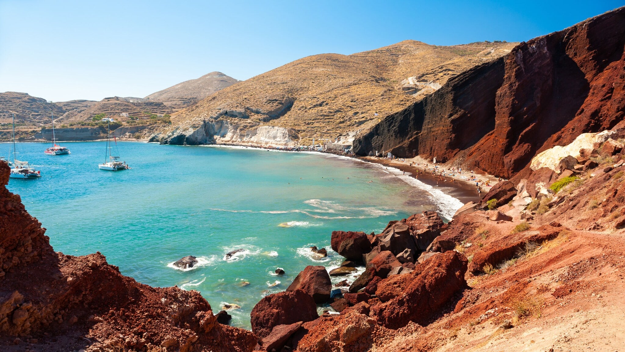 Meeresbucht mit Booten im türkisblauen Wasser, umgeben von roten Felsklippen an rotem, belebtem Sandstrand. Meeresbucht mit Booten im türkisblauen Wasser, umgeben von roten Felsklippen an rotem, belebtem Sandstrand.