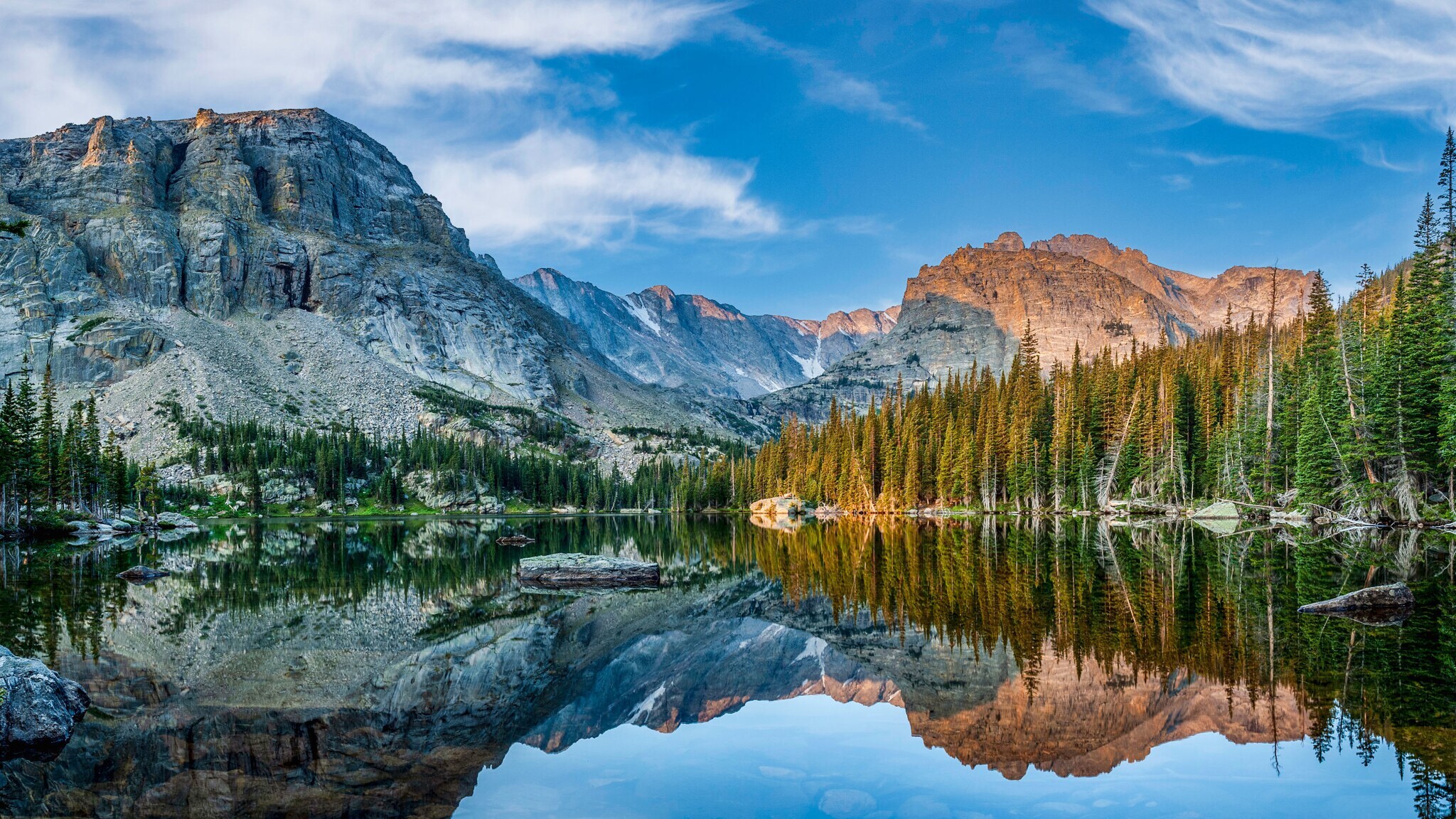 Idyllische Berglandschaft mit Nadelwald vor schroffen Felsen, die sich in einem See spiegeln unter blauem Himmel.