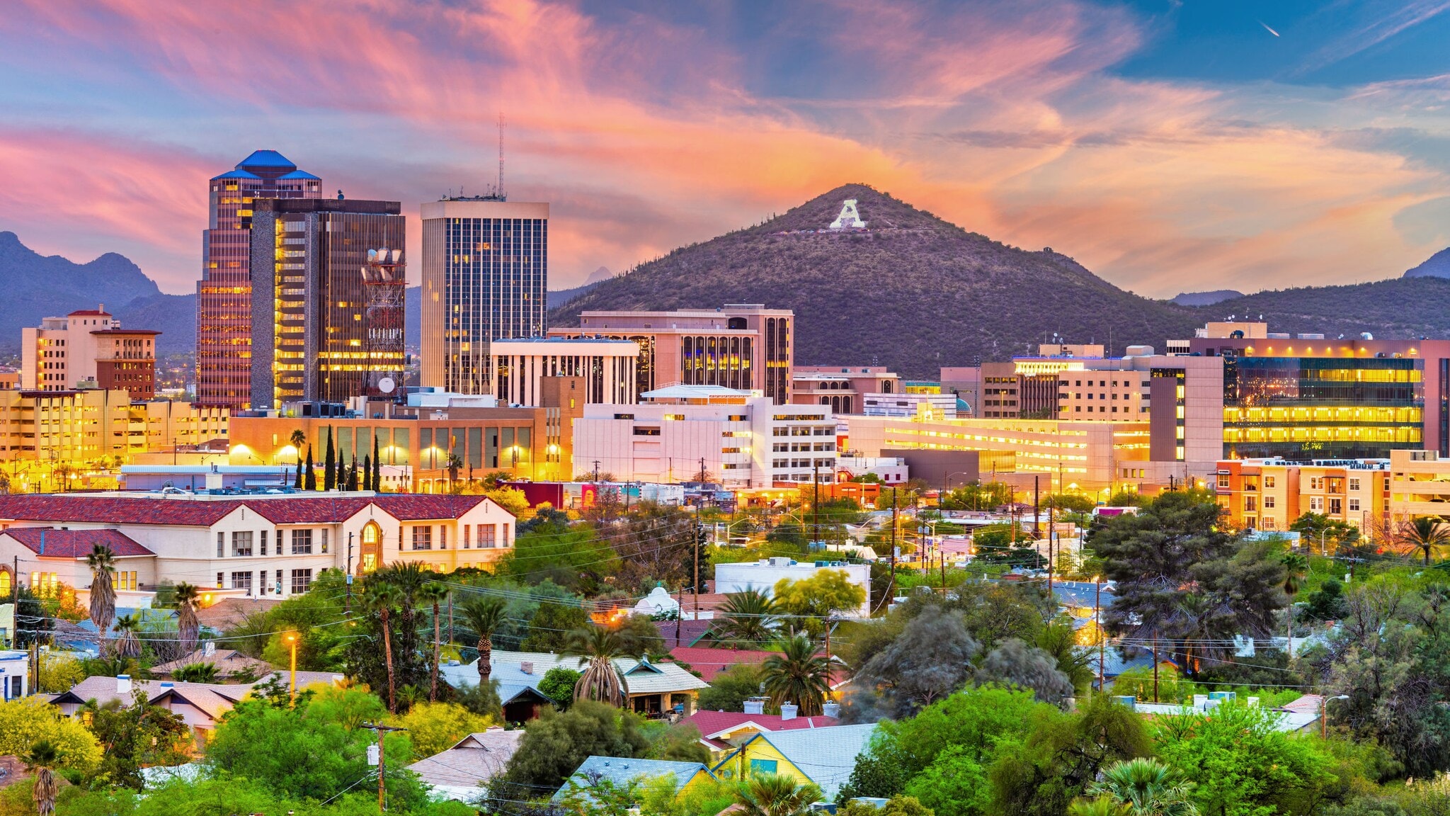 Beleuchte Skyline von Tucson mit Hochhäusern und grüner Vegetation vor Gebirgszug bei Sonnenuntergang. Beleuchte Skyline von Tucson mit Hochhäusern und grüner Vegetation vor Gebirgszug bei Sonnenuntergang.