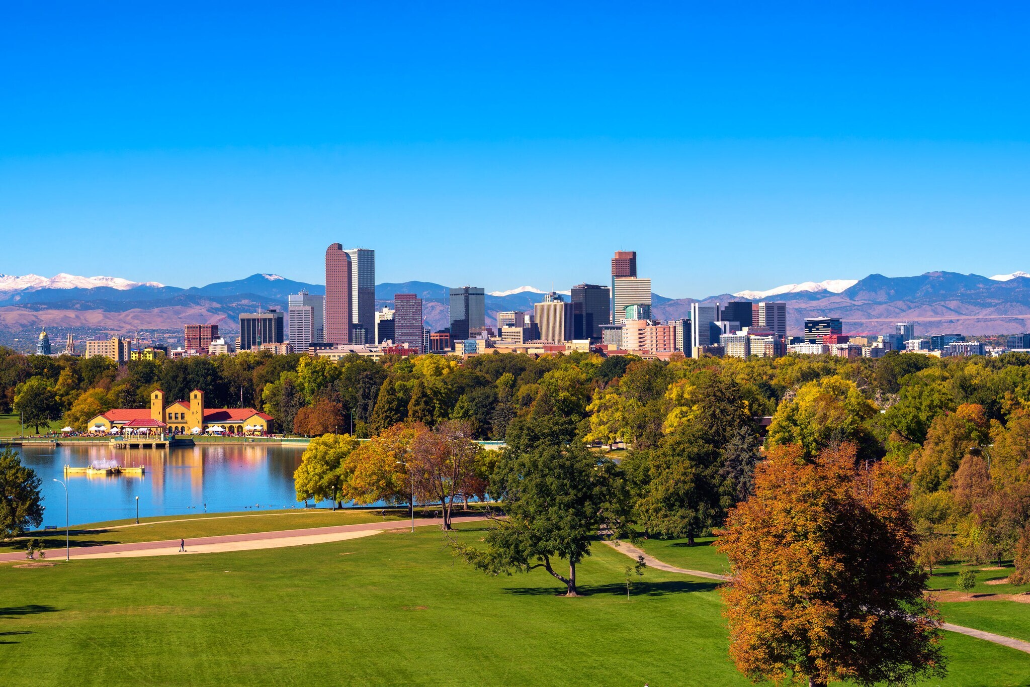 Skyline von Denver unter blauem Himmel vor Gebirgszug, im Vordergrund eine Parkanlage mit Teich und grünen Bäumen.