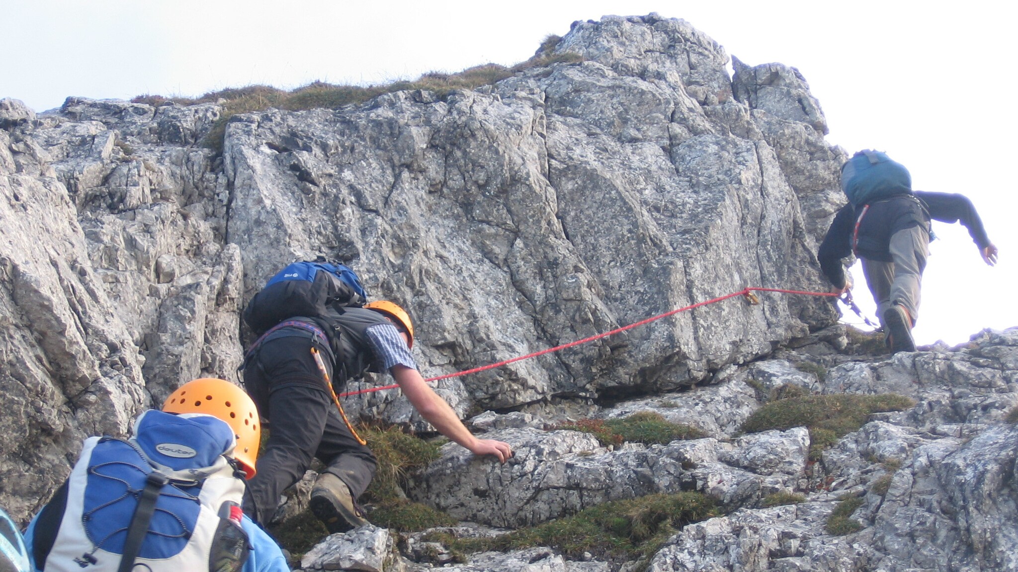 Drei Personen mit Kletterausrüstung, die einen steilen Felsen hochklettern, an dem ein Seil gespannt ist.