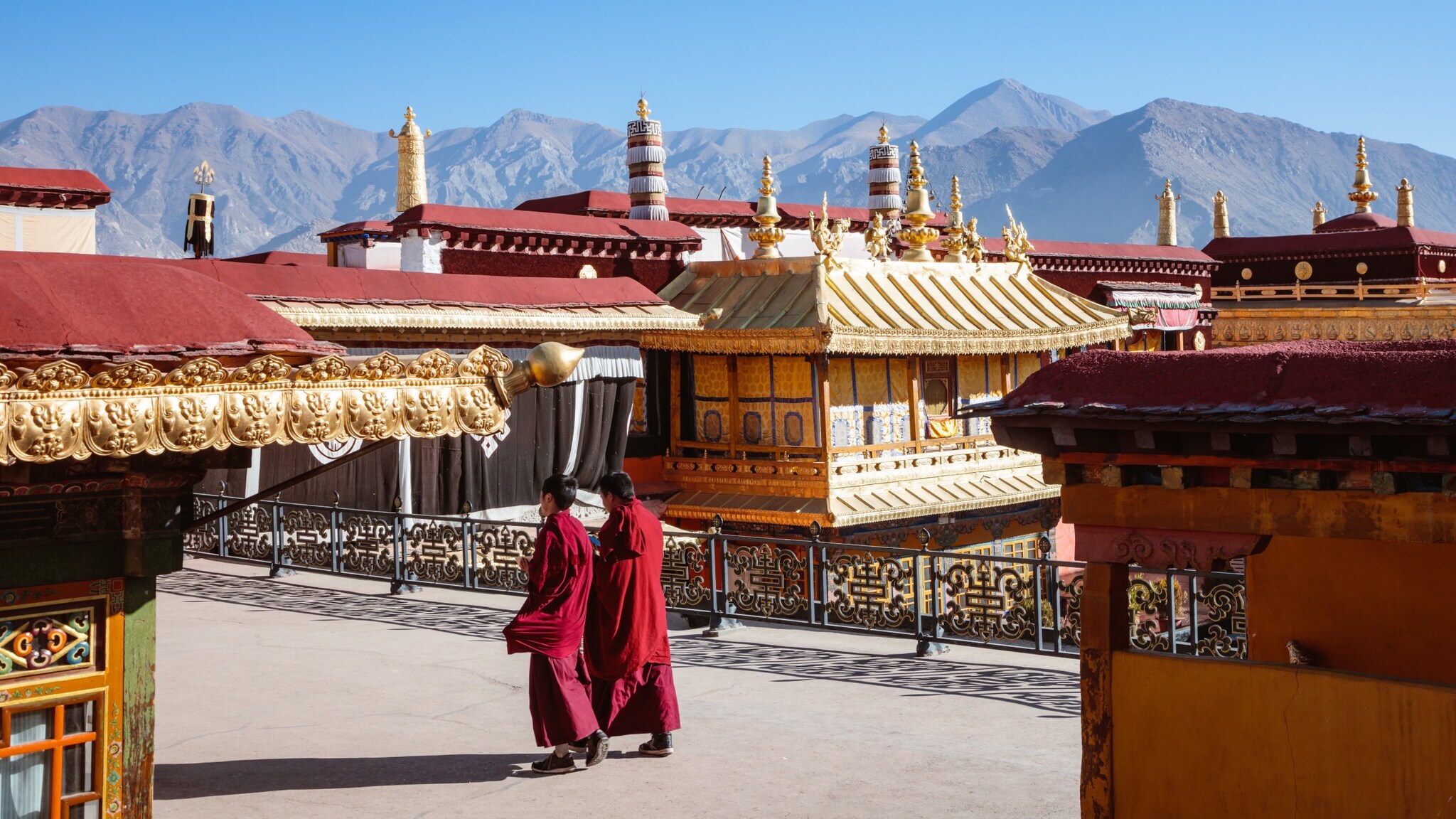 Zwei tibetische Mönche in roten Kutten gehen durch eine buddhistische Tempelanlage vor Bergpanorama.