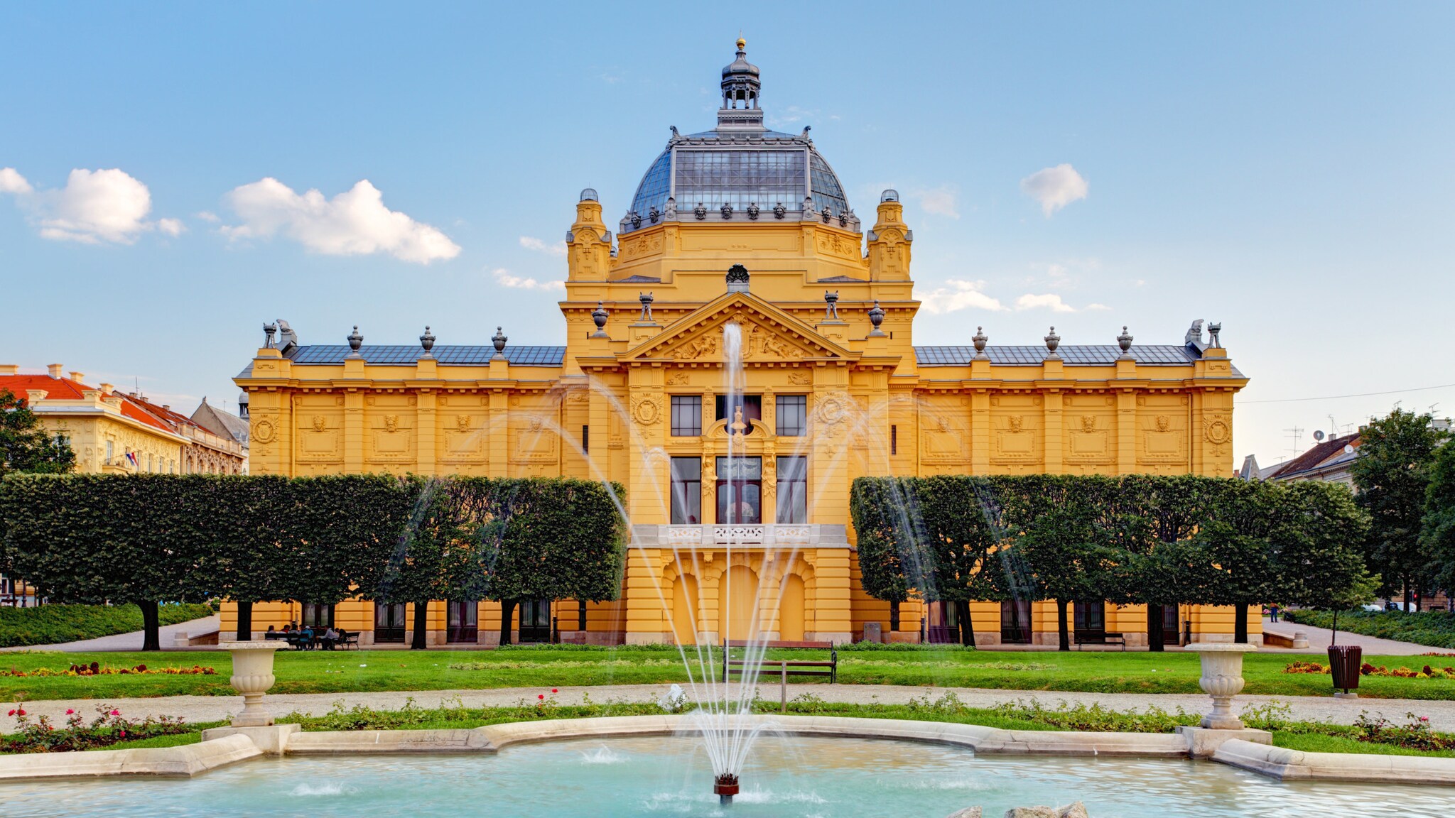 Gelbes Jugendstilgebäude mit Glaskuppel in einer Parkanlage mit Springbrunnen im Vordergrund.