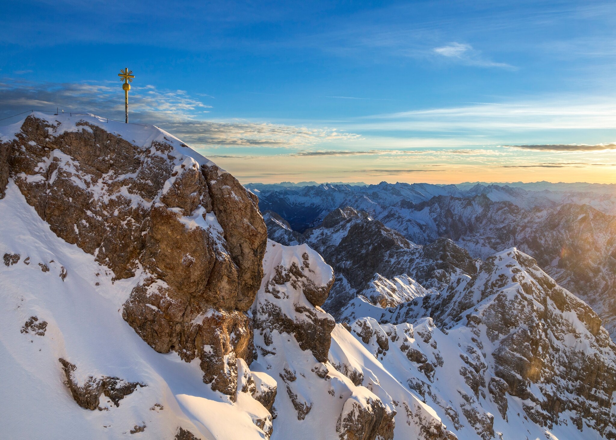 Blick auf den teilweise verschneiten Gipfel der Zugspitze mit Gipfelkreuz, im Hintergrund Berge und blauer Himmel. Der anspruchsvolle Aufstieg über den Höllental-Klettersteig auf die Zugspitze belohnt mit einer Top-Aussicht.
