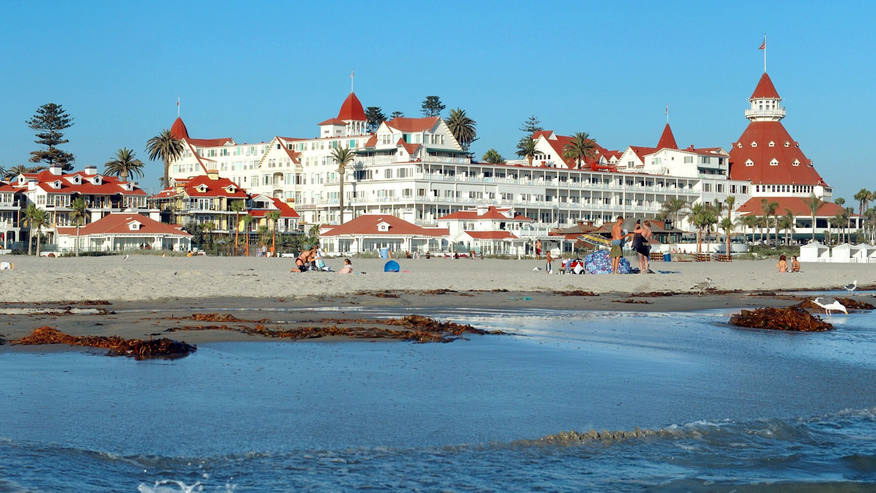 Blick vom Wasser auf einen Sandstrand mit Palmen und Personen vor einem weißen Hotelkomplex im viktorianischen Stil.