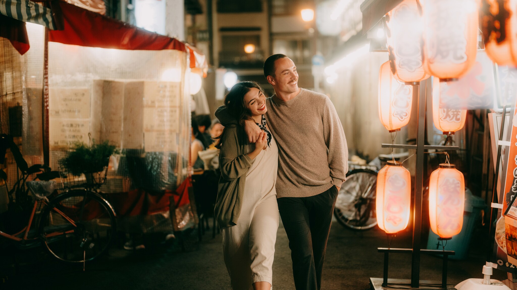 Ein Paar läuft Arm in Arm eine beleuchtete Gasse mit asiatischen Lampions am Abend entlang. Ein Paar läuft Arm in Arm eine beleuchtete Gasse mit asiatischen Lampions am Abend entlang.