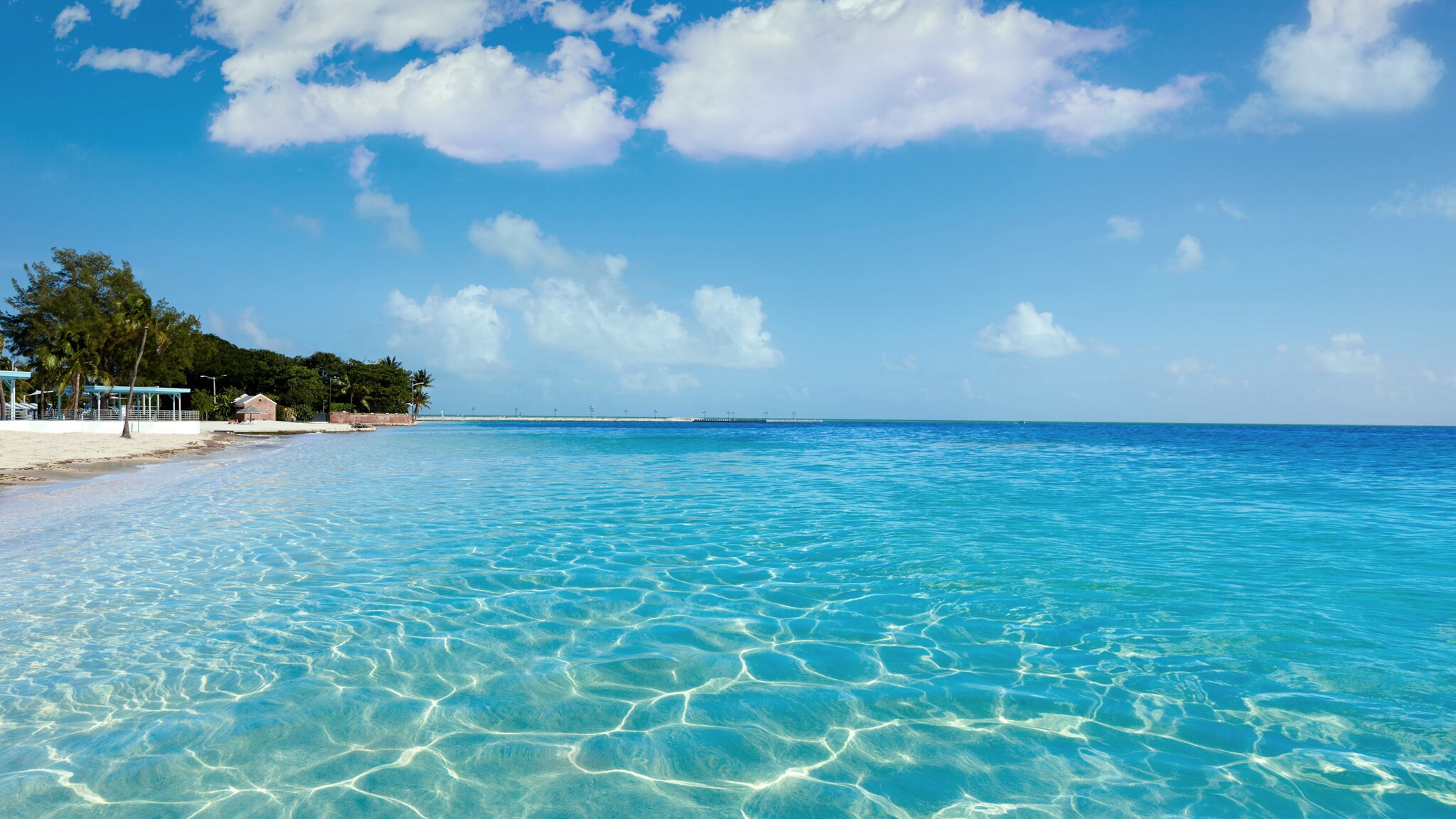 Flaches türkisblaues Meer vor einem weißen Sandstrand unter blauem Himmel.