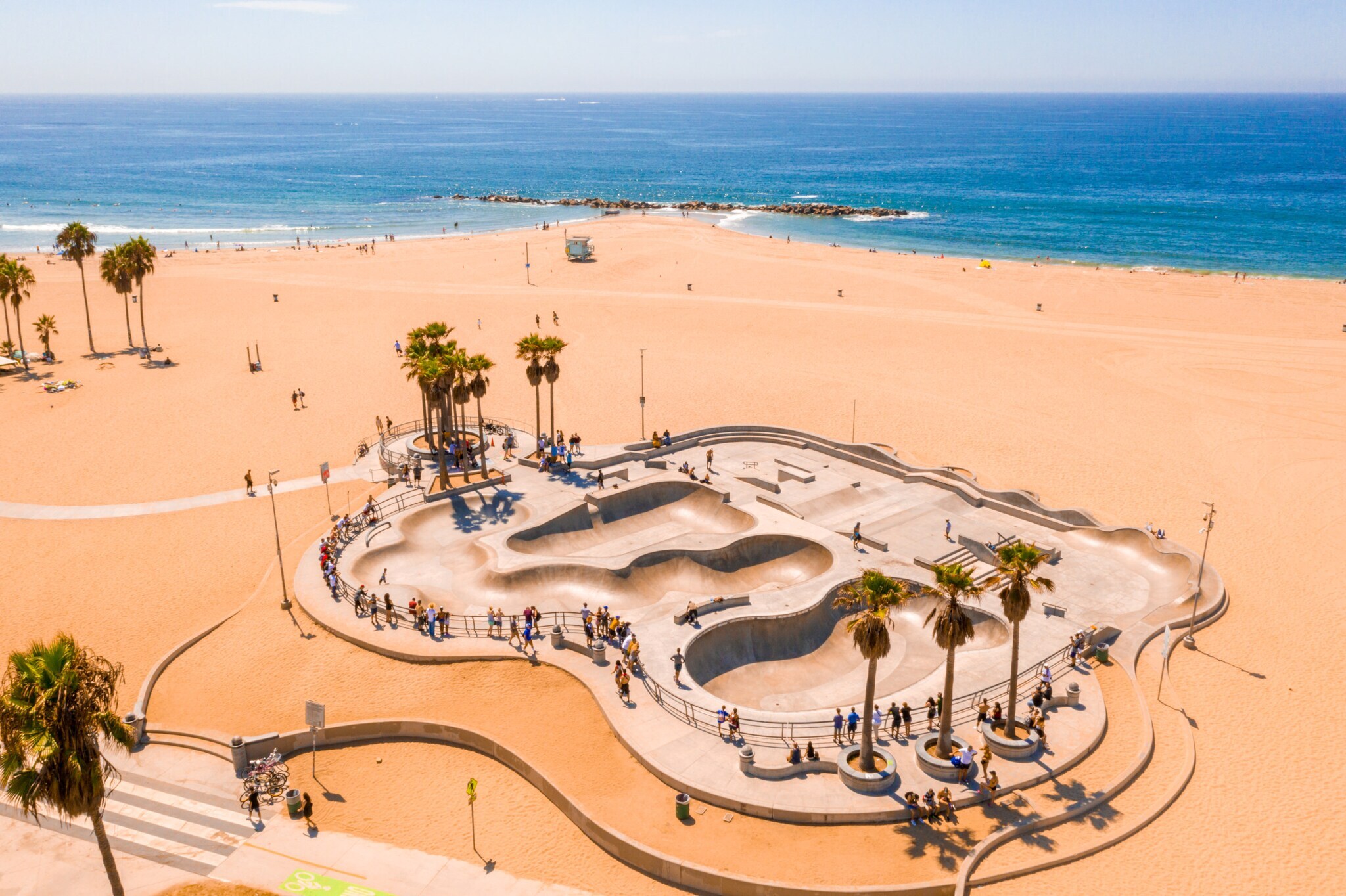 Personen an einem Skatepark mit Betonrampen und Palmen an einem breiten Sandstrand.