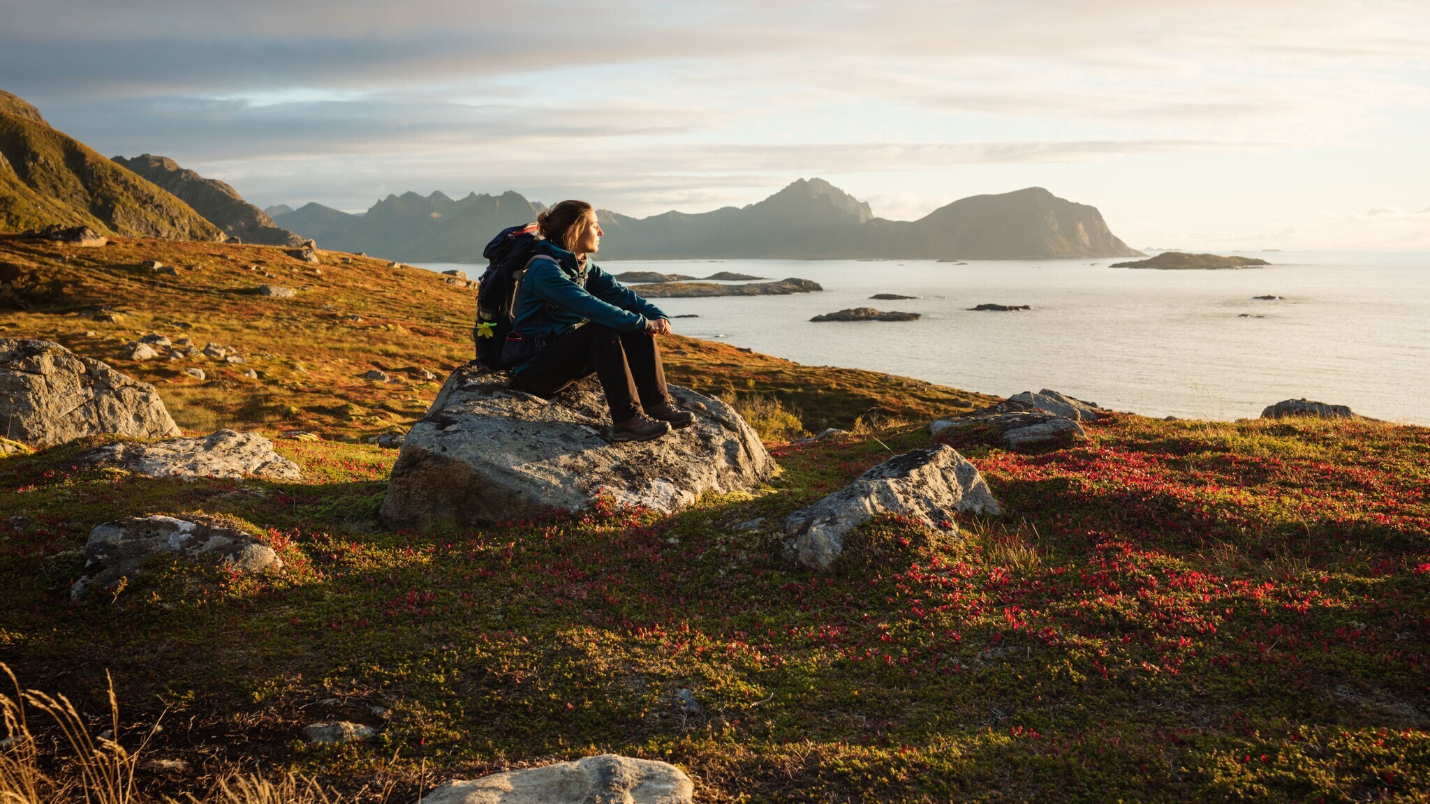 Eine Frau mit Wanderausrüstung, die bei Sonnenuntergang auf einem großen Stein sitzt und aufs Wasser blickt, im Hintergrund Berge. Eine Frau mit Wanderausrüstung, die bei Sonnenuntergang auf einem großen Stein sitzt und aufs Wasser blickt, im Hintergrund Berge.