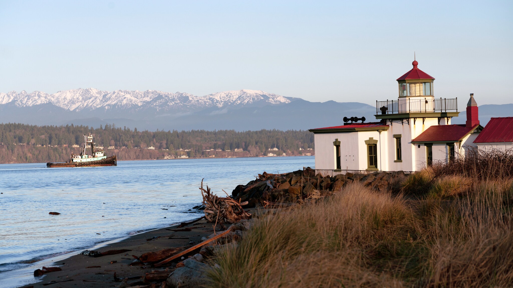 Niedriger Leuchtturm auf einem kleinen Sandstrand mit Dünen an einer Meerenge mit einem Kutter vor schneebedeckter Gebirgskette. Niedriger Leuchtturm auf einem kleinen Sandstrand mit Dünen an einer Meerenge mit einem Kutter vor schneebedeckter Gebirgskette.