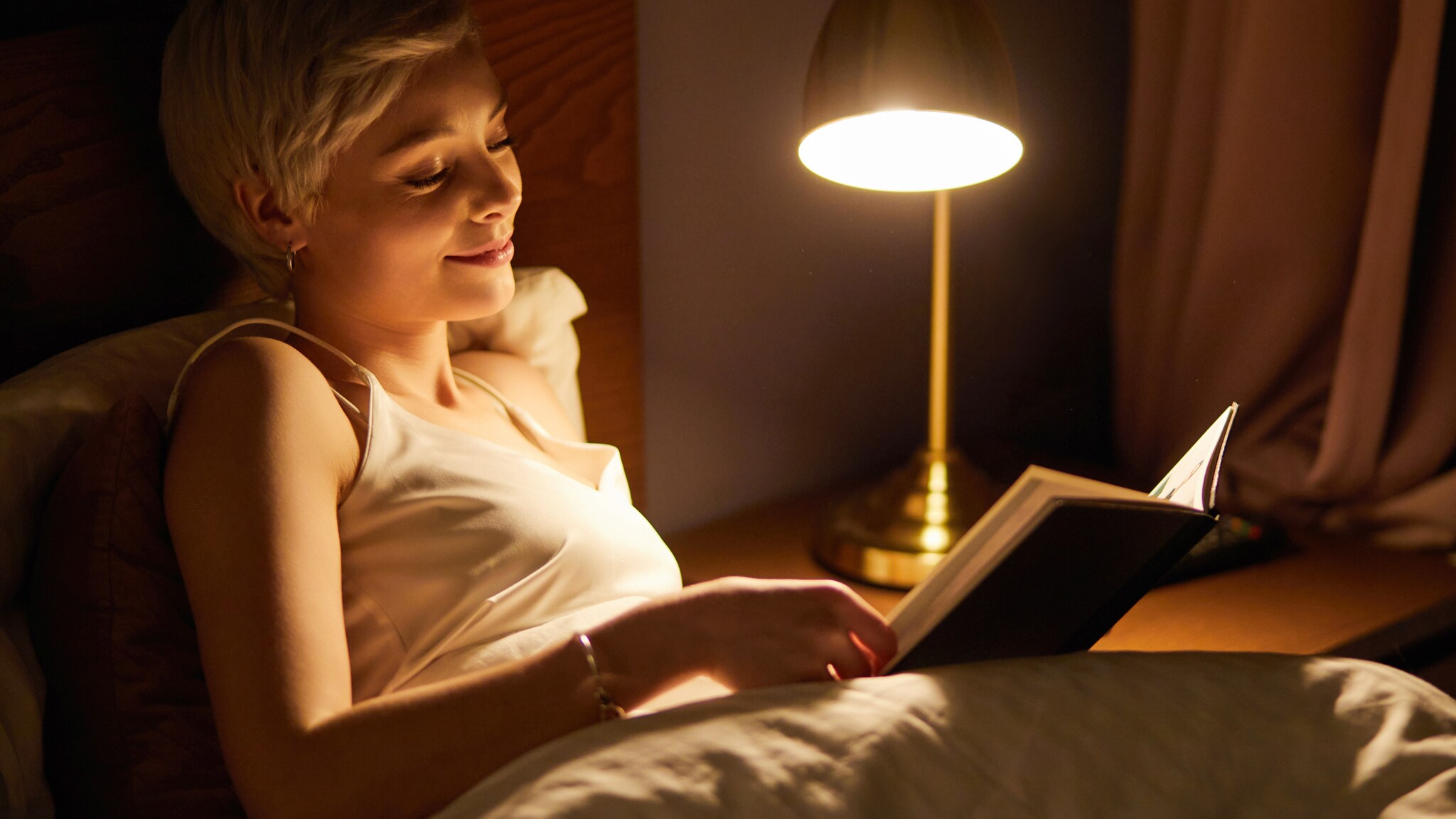 Eine Frau liest im Bett ein Buch im Licht einer Nachttischlampe.