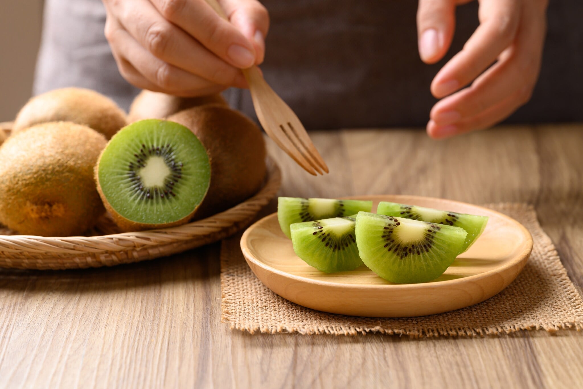 Nahaufnahme von geschnittenen Kiwis auf einem Holzteller, darüber zwei Hände mit Holzgabel.