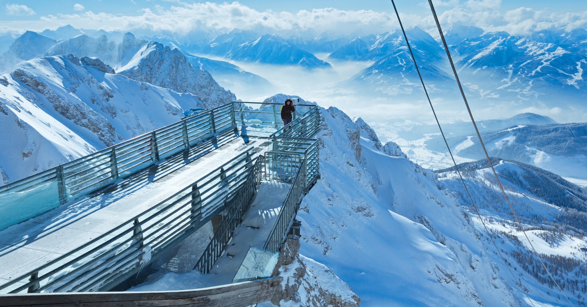 Eine Person steht auf einer Aussichtsplattform vor schneebedecktem Bergpanorama.