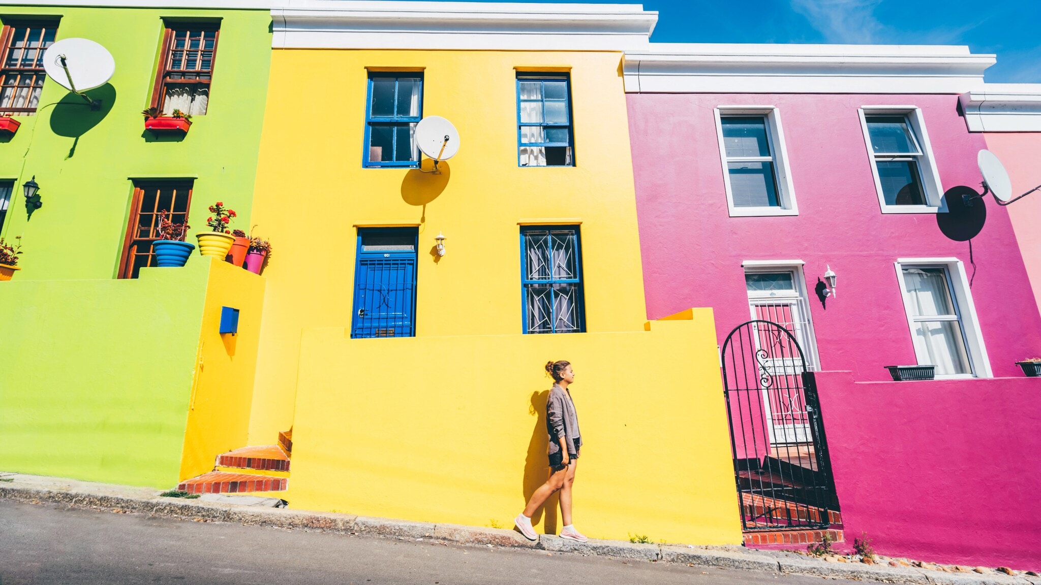 Eine Person geht vor einer Häuserfront bestehend aus Häusern in unterschiedlichen knalligen Farben bei Sonnenschein. Eine Person geht vor einer Häuserfront bestehend aus Häusern in unterschiedlichen knalligen Farben bei Sonnenschein.