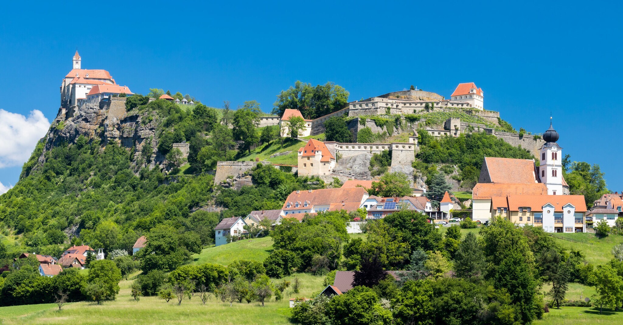 Eine Burganlage mit mehreren Gebäuden auf einem begrünten Felsen unter blauem Himmel.