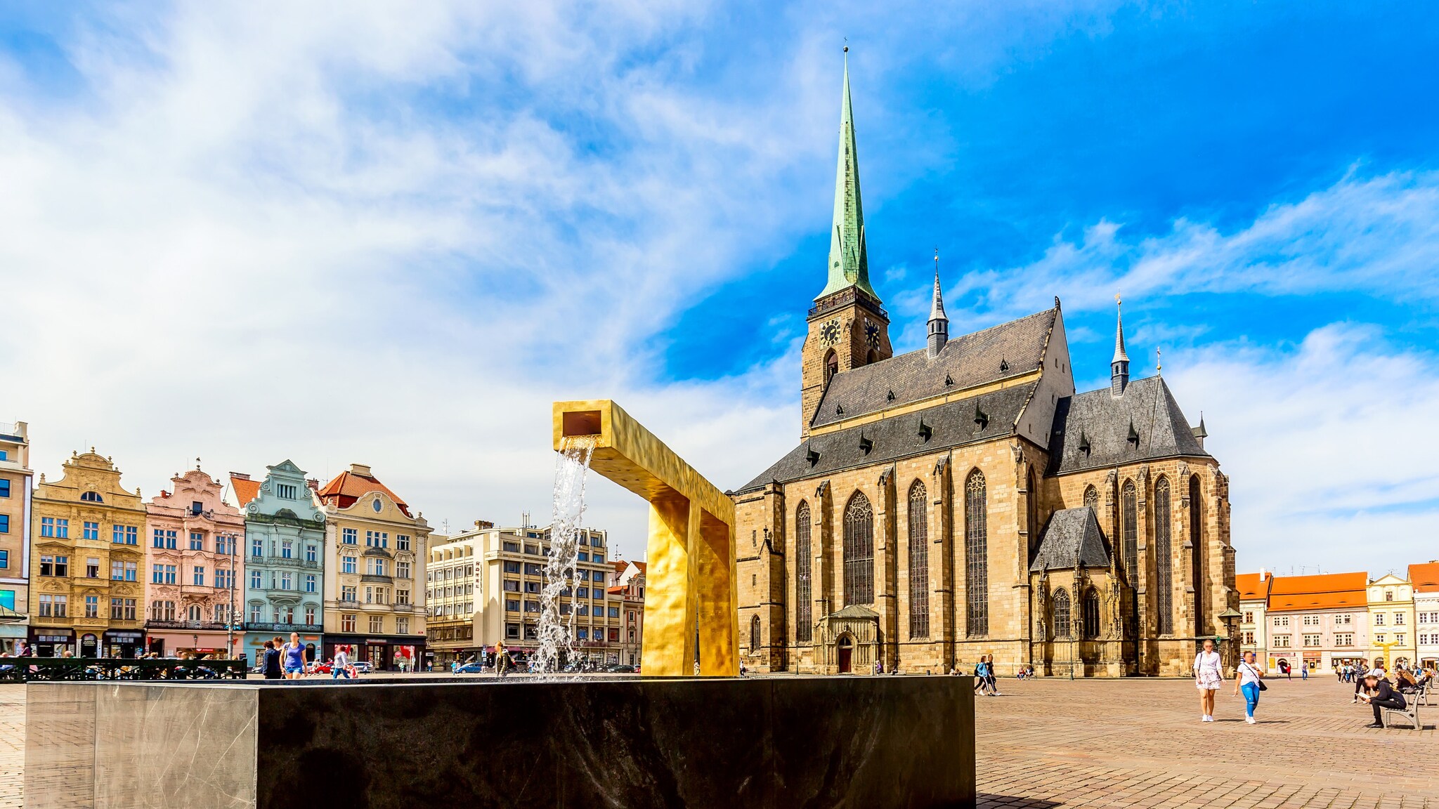 Ein moderner Springbrunnen auf einem Platz mit bunten, historischen Häusern vor einer Kathedrale.