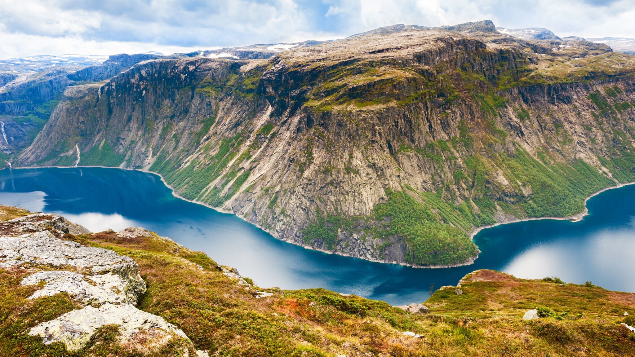 Eisblaues Wasser eines Fjords in Norwegen mit Klippe im Vordergrund und Bergen im Hintergrund.