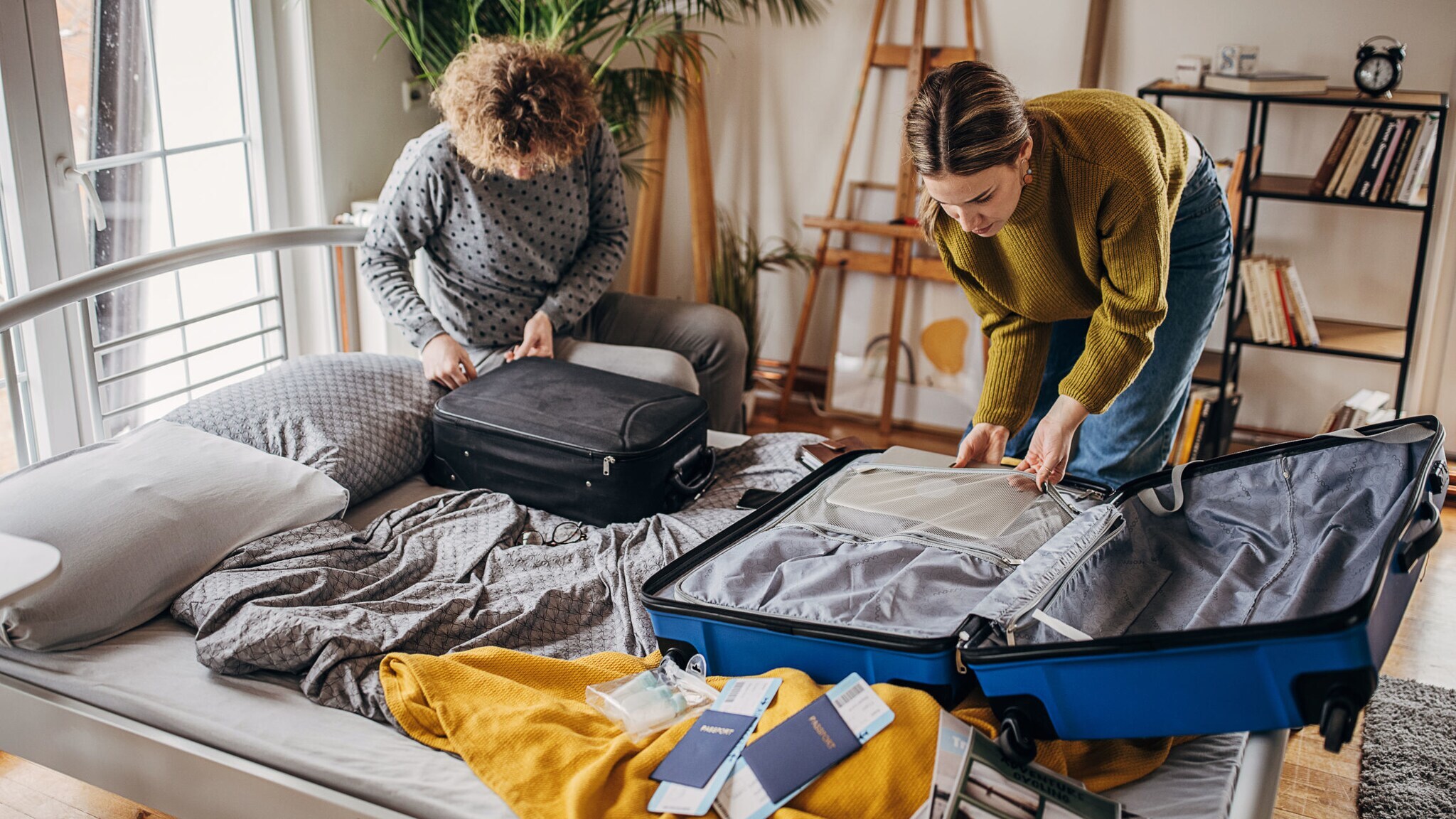 Eine Frau und ein Mann packen gemeinsam jeweils einen Koffer auf einem Bett, Reisepapiere liegen daneben.