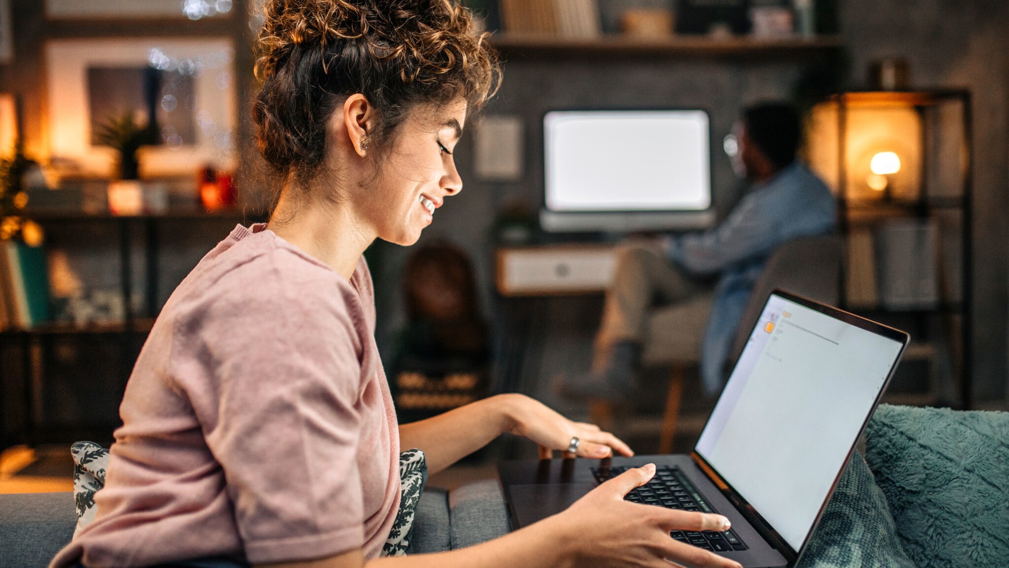 Eine Frau sitzt mit aufgeklapptem Laptop auf einer Couch, im Hintergrund sitzt ein Mann an einem Schreibtisch mit PC. Eine Frau sitzt mit aufgeklapptem Laptop auf einer Couch, im Hintergrund sitzt ein Mann an einem Schreibtisch mit PC.