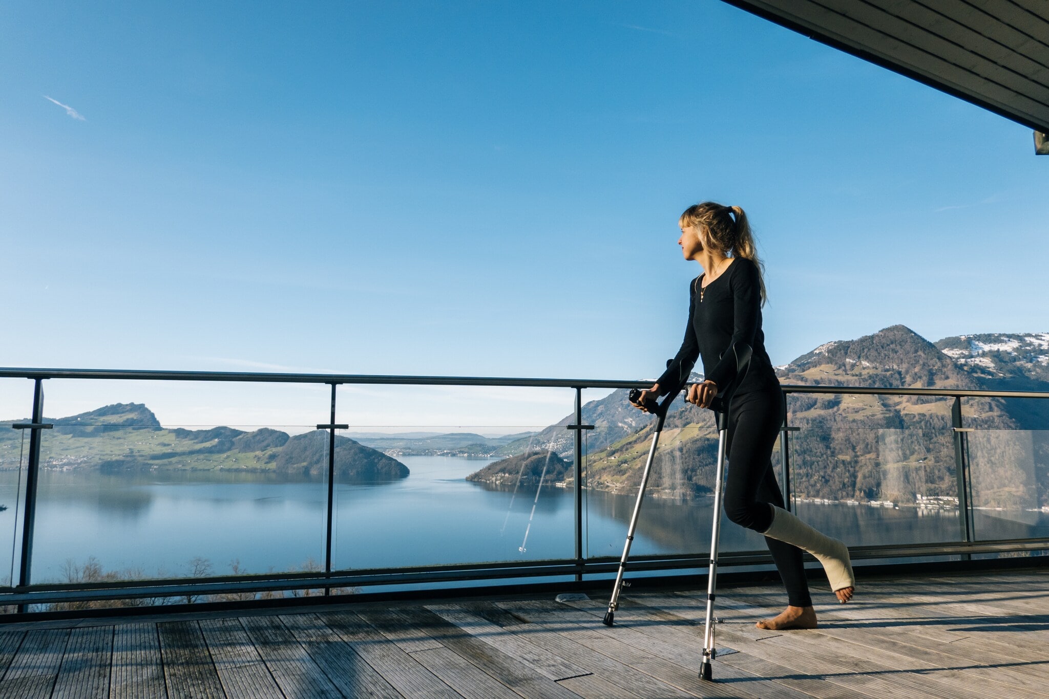 Eine Frau mit eingegipstem Bein steht mit Krücken auf einer Terrasse mit Blick auf eine Seenlandschaft mit bergigem Umland. Eine Frau mit eingegipstem Bein steht mit Krücken auf einer Terrasse mit Blick auf eine Seenlandschaft mit bergigem Umland.