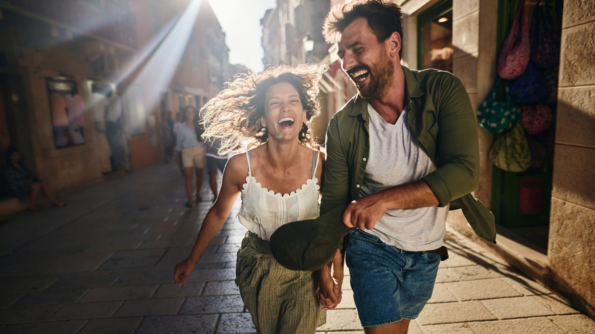 Ein lachendes Paar läuft durch eine Einkaufsstraße in einem mediterranen Ort bei Sonnenschein.