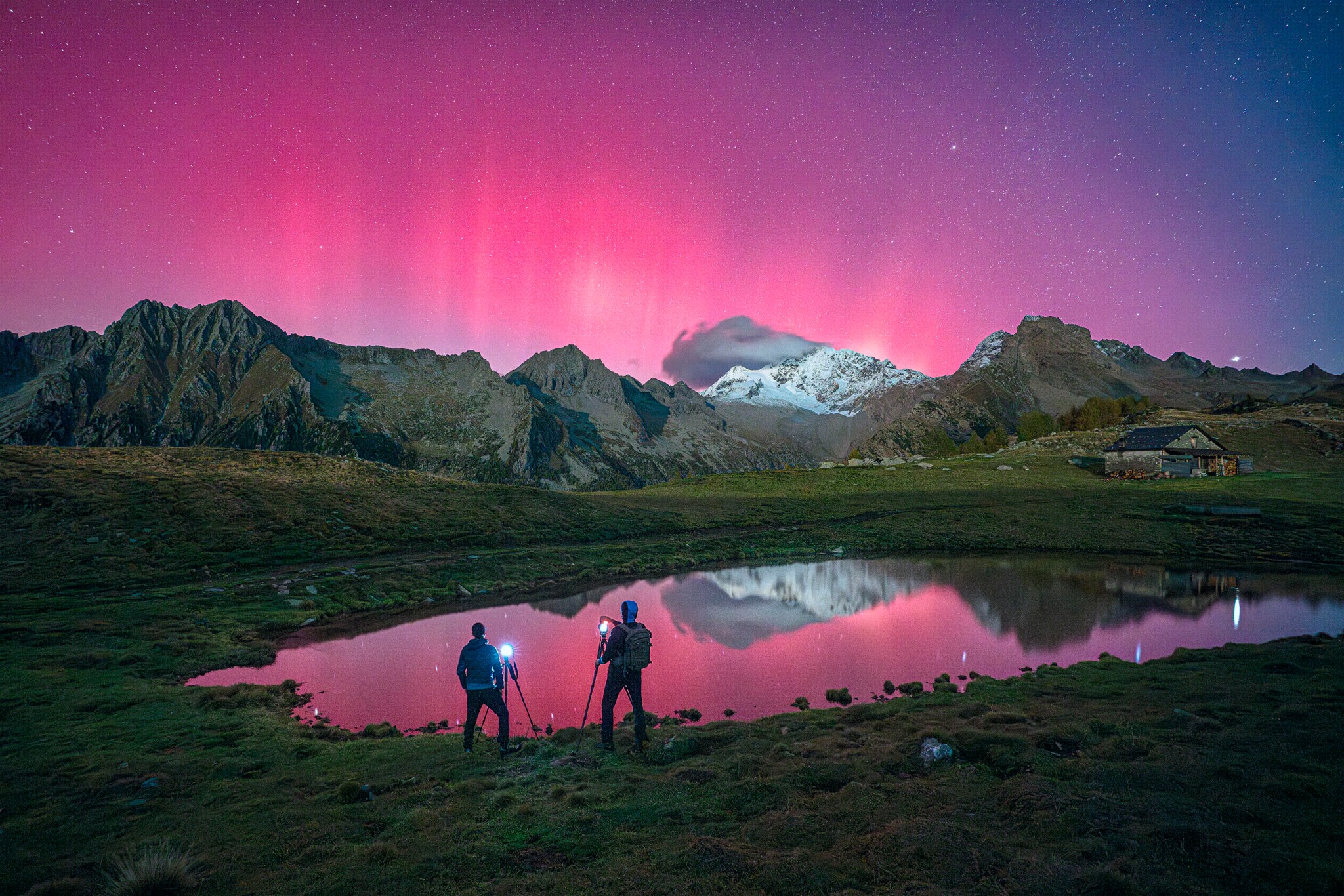 Zwei Menschen stehen vor einer Bergkulisse und fotografieren Polarlichter, die den Himmel pink färben. Zwei Menschen stehen vor einer Bergkulisse und fotografieren Polarlichter, die den Himmel pink färben.