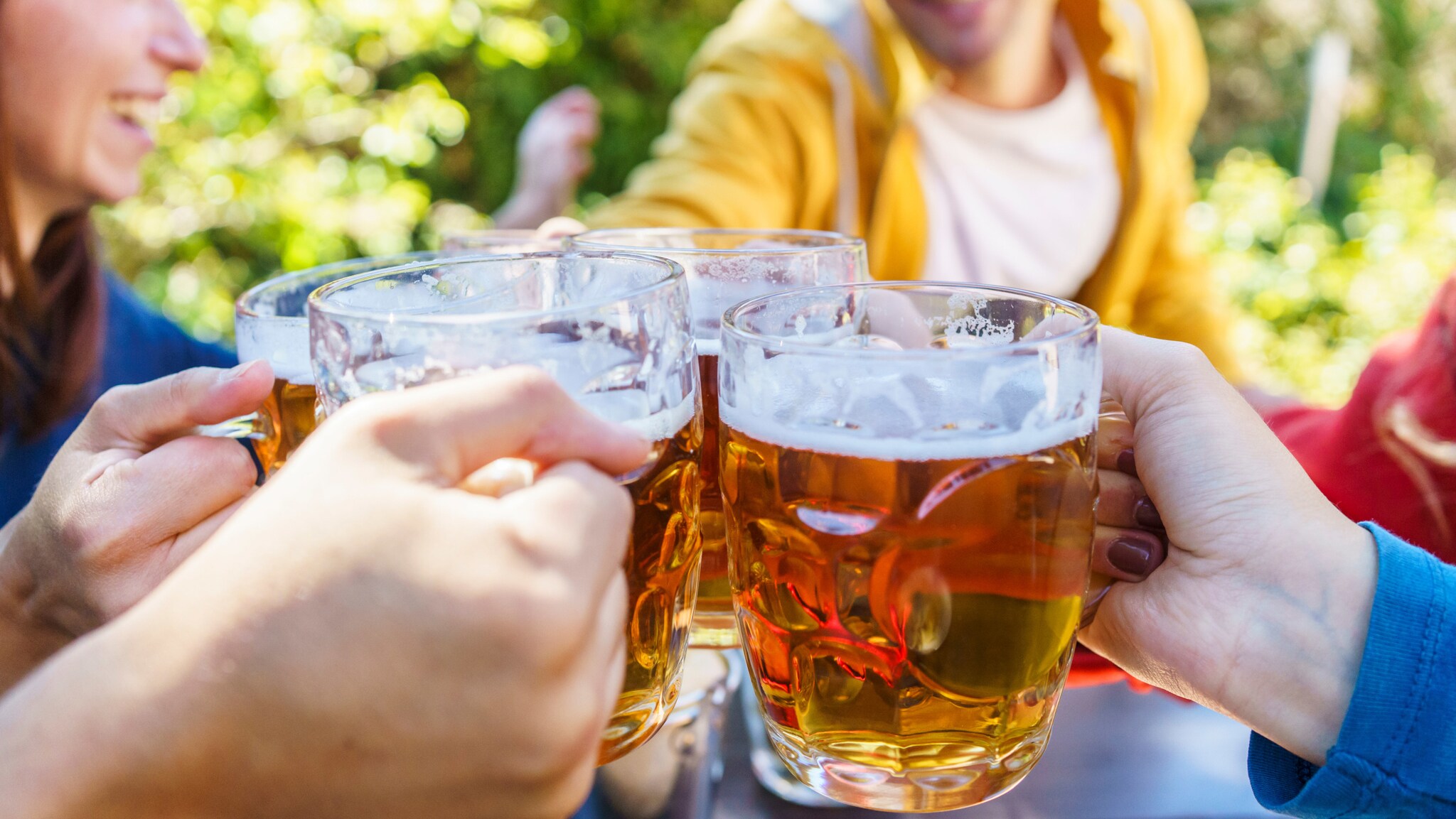 Nahaufnahme von vier gefüllten Bierkrügen aus Glas in den Händen von vier Personen beim Anstoßen an einem Tisch im Freien.
