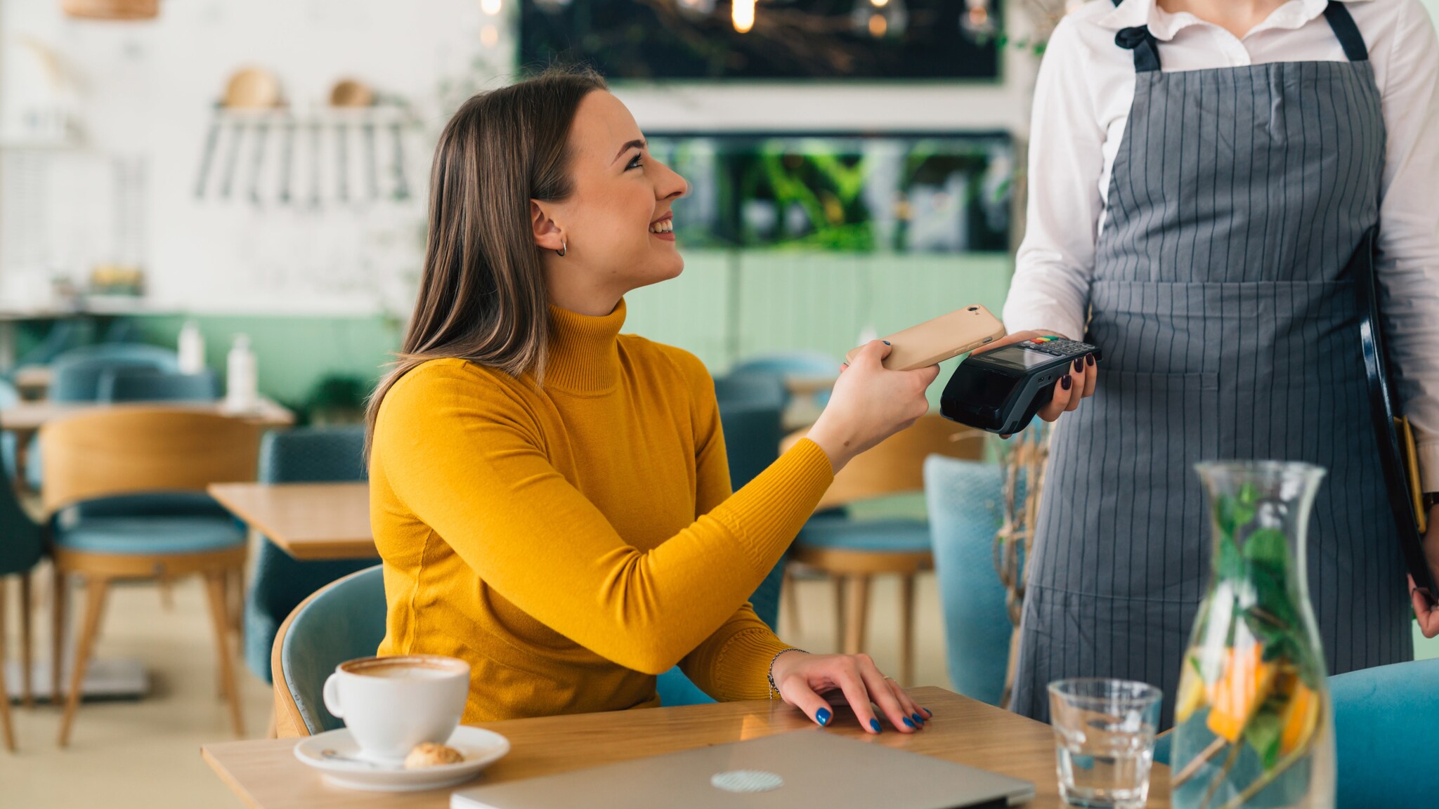 Eine Frau bezahlt kontaktlos mit ihrem Smartphone an einem Tisch in einem Café.