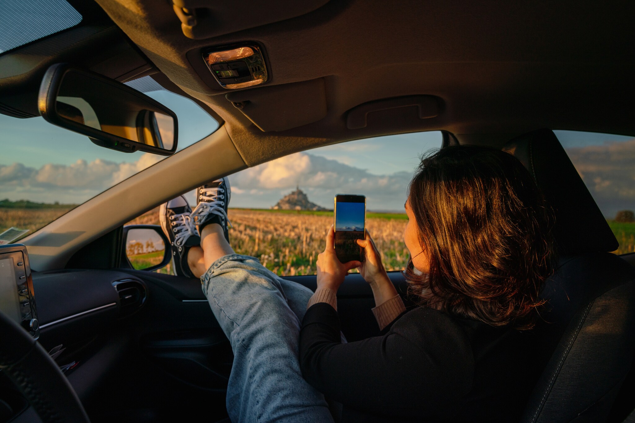 Eine Frau auf dem Beifahrersitz eines Autos hält die Beine aus dem Fenster und fotografiert die Landschaft mit dem Smartphone. Eine Frau auf dem Beifahrersitz eines Autos hält die Beine aus dem Fenster und fotografiert die Landschaft mit dem Smartphone.