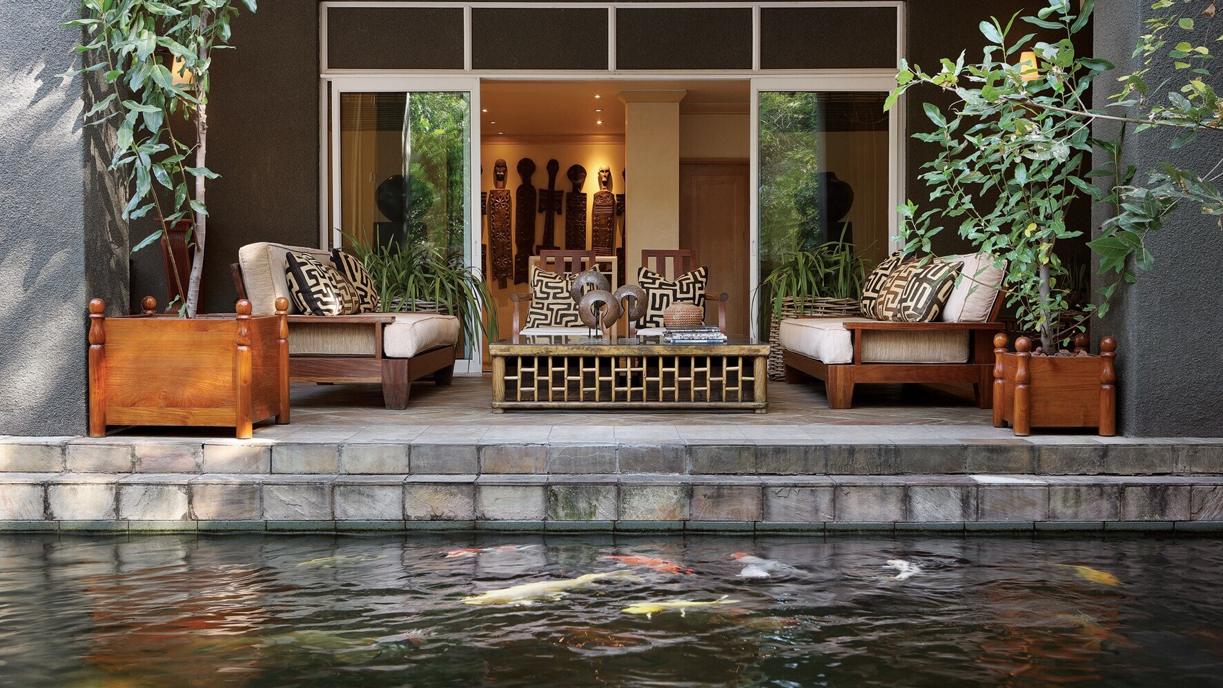 Terrasse mit Holzmöbeln und Kunstwerken im afrikanischen Stil an einem Wasserbecken mit Koikarpfen. Terrasse mit Holzmöbeln und Kunstwerken im afrikanischen Stil an einem Wasserbecken mit Koikarpfen.