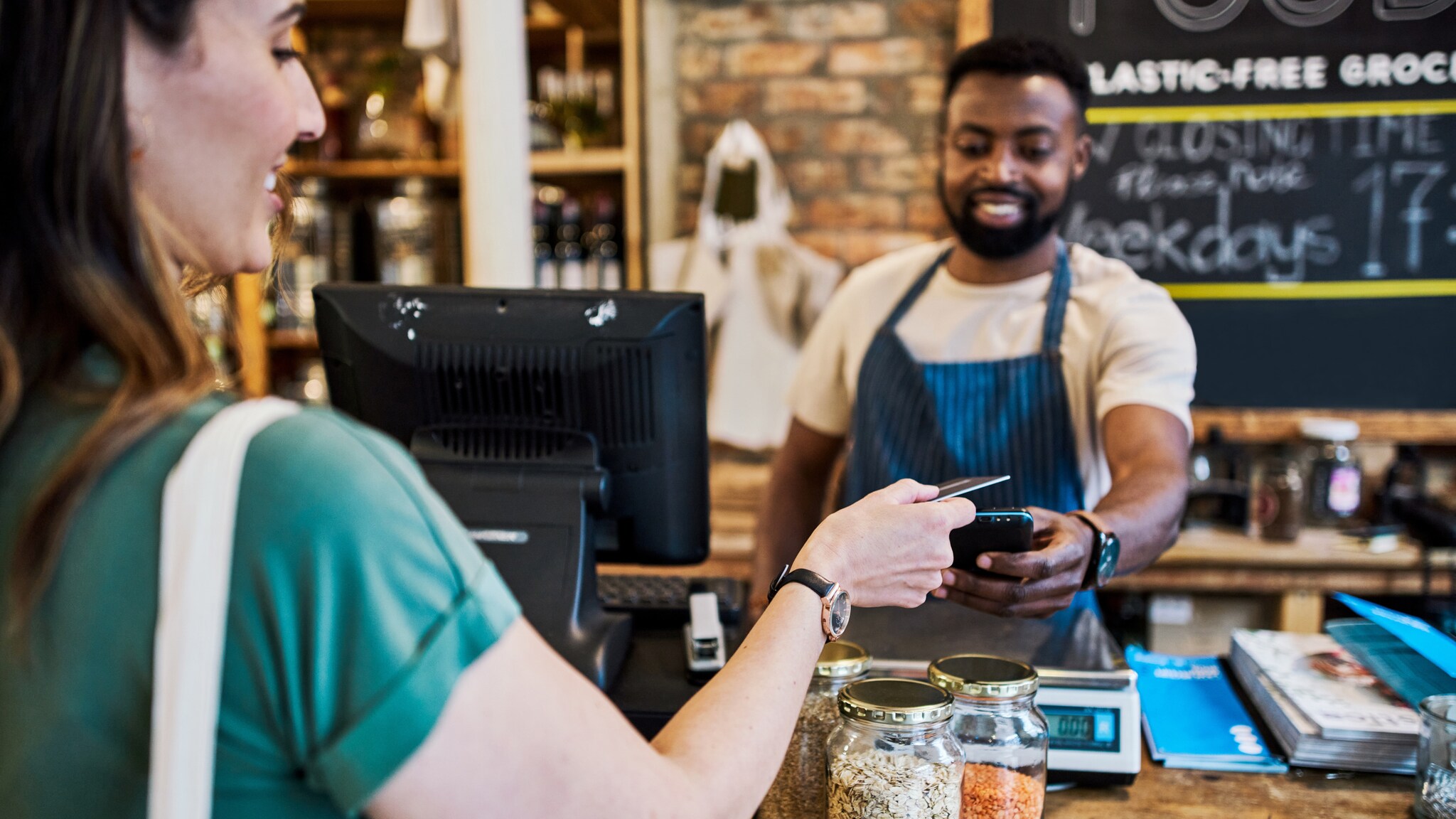 Eine Frau bezahlt kontaktlos mit ihrer Kreditkarte an der Theke in einem Geschäft, der Verkäufer hält ihr ein Kartenlesegerät entgegen. Eine Frau bezahlt kontaktlos mit ihrer Kreditkarte an der Theke in einem Geschäft, der Verkäufer hält ihr ein Kartenlesegerät entgegen.