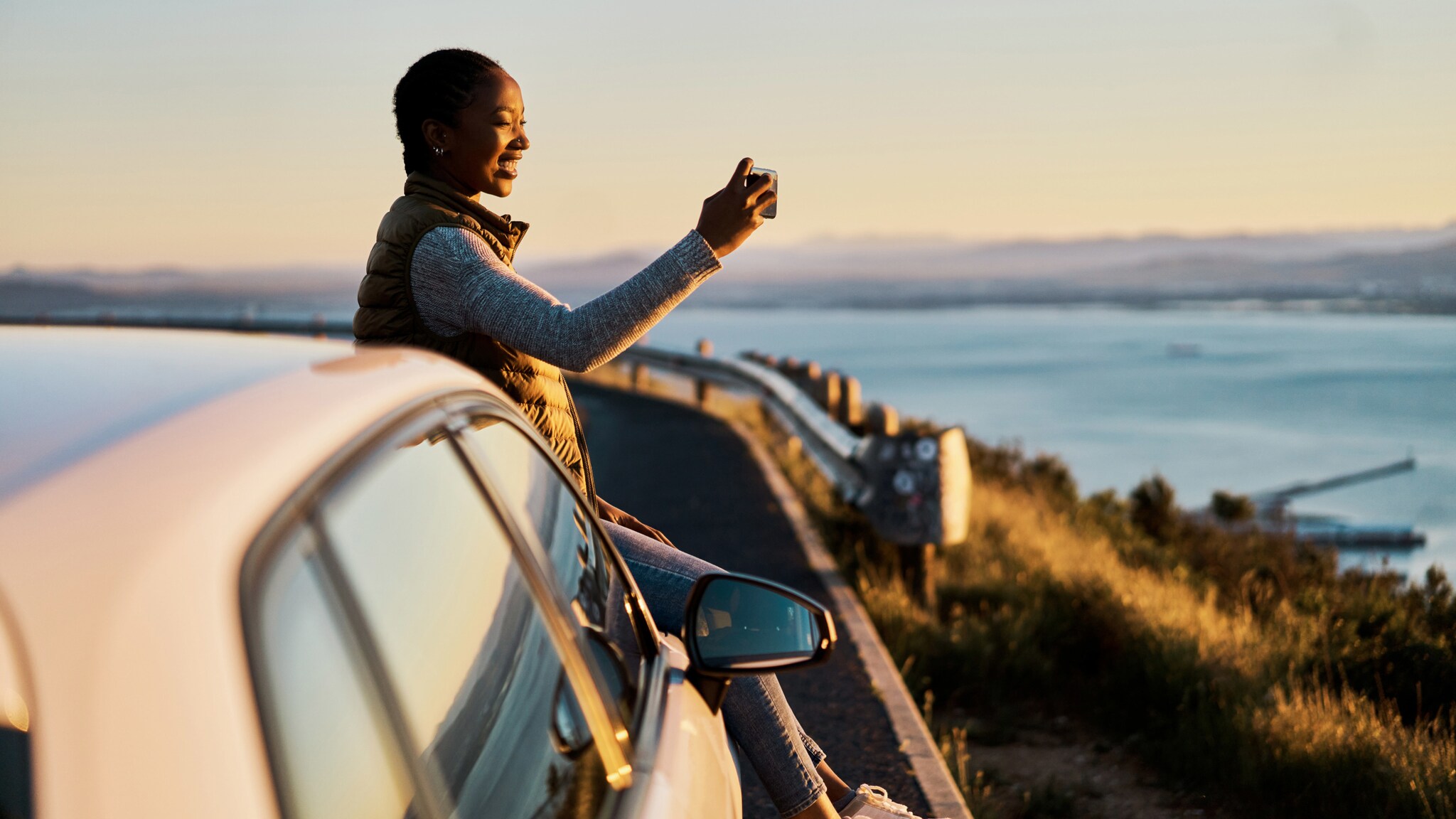 Eine Frau sitzt auf der Motorhaube eines parkenden Autos auf einer Küstenstraße und macht ein Foto mit ihrem Smartphone.