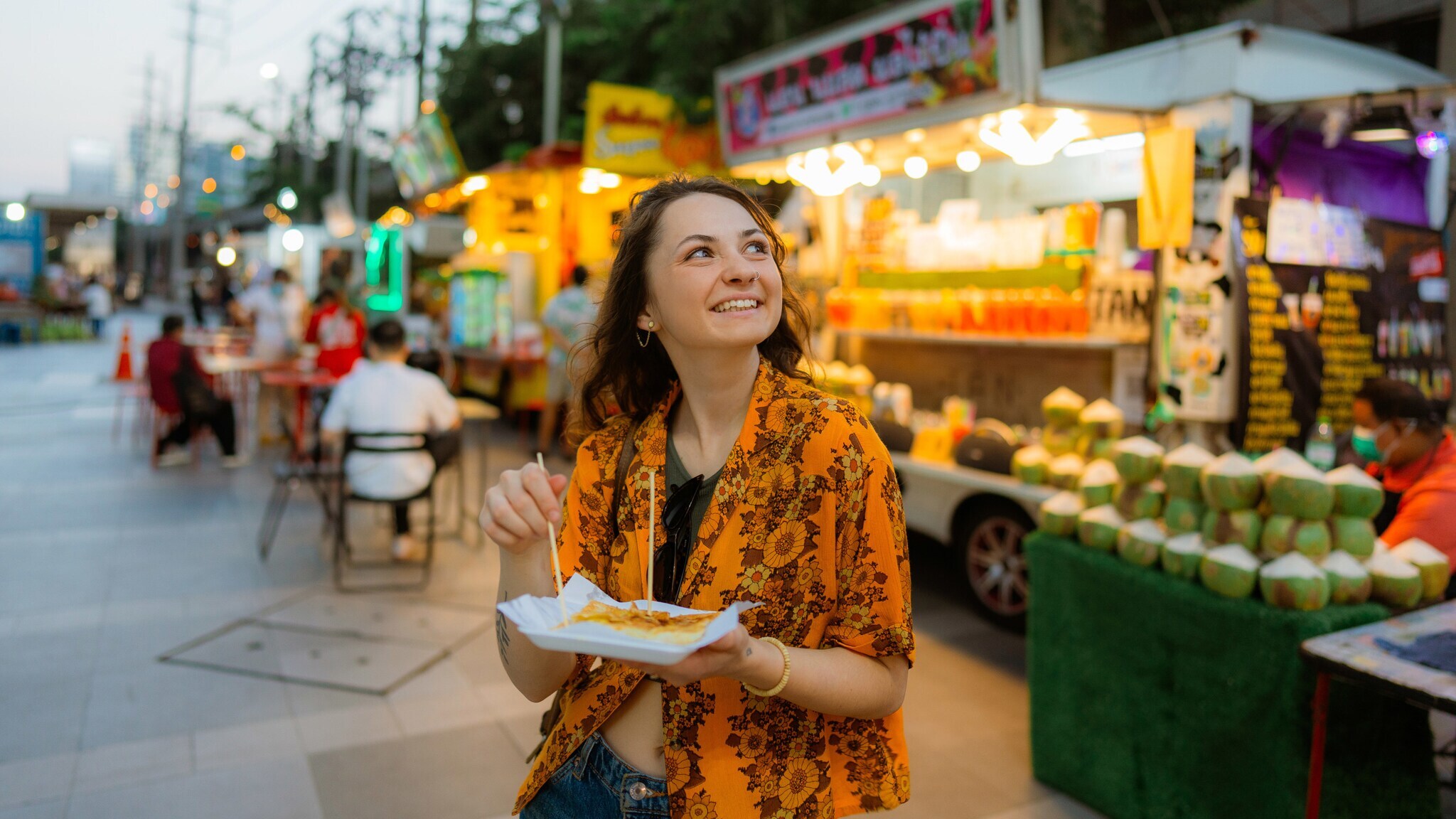 Eine junge Frau mit einer Speise in ihrer Hand vor einem asiatischen Streetfood-Stand.
