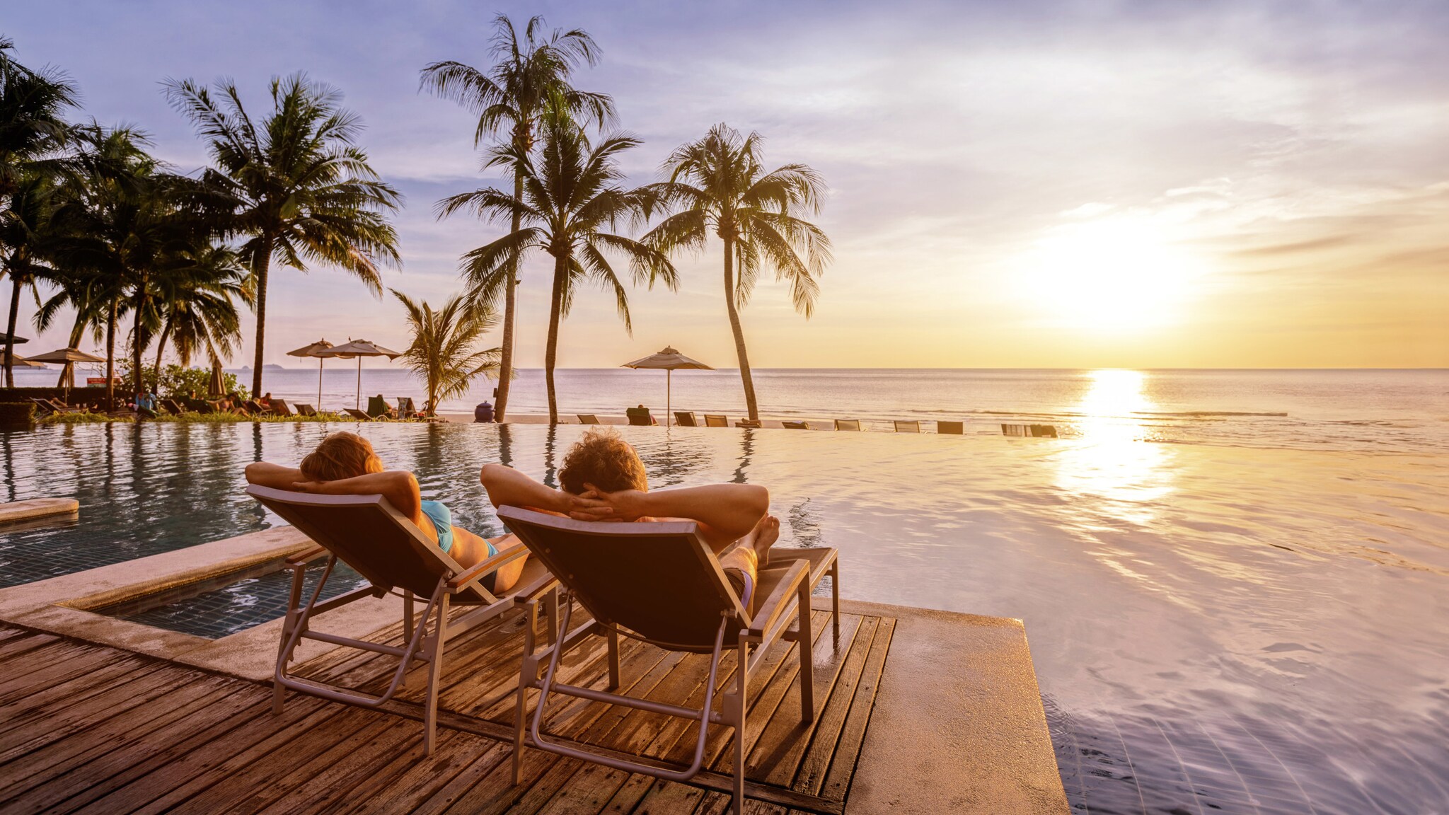 Rückansicht eines Paares in Liegestühlen an einem Hotelpool mit Palmen am Meer bei Sonnenuntergang. Rückansicht eines Paares in Liegestühlen an einem Hotelpool mit Palmen am Meer bei Sonnenuntergang.