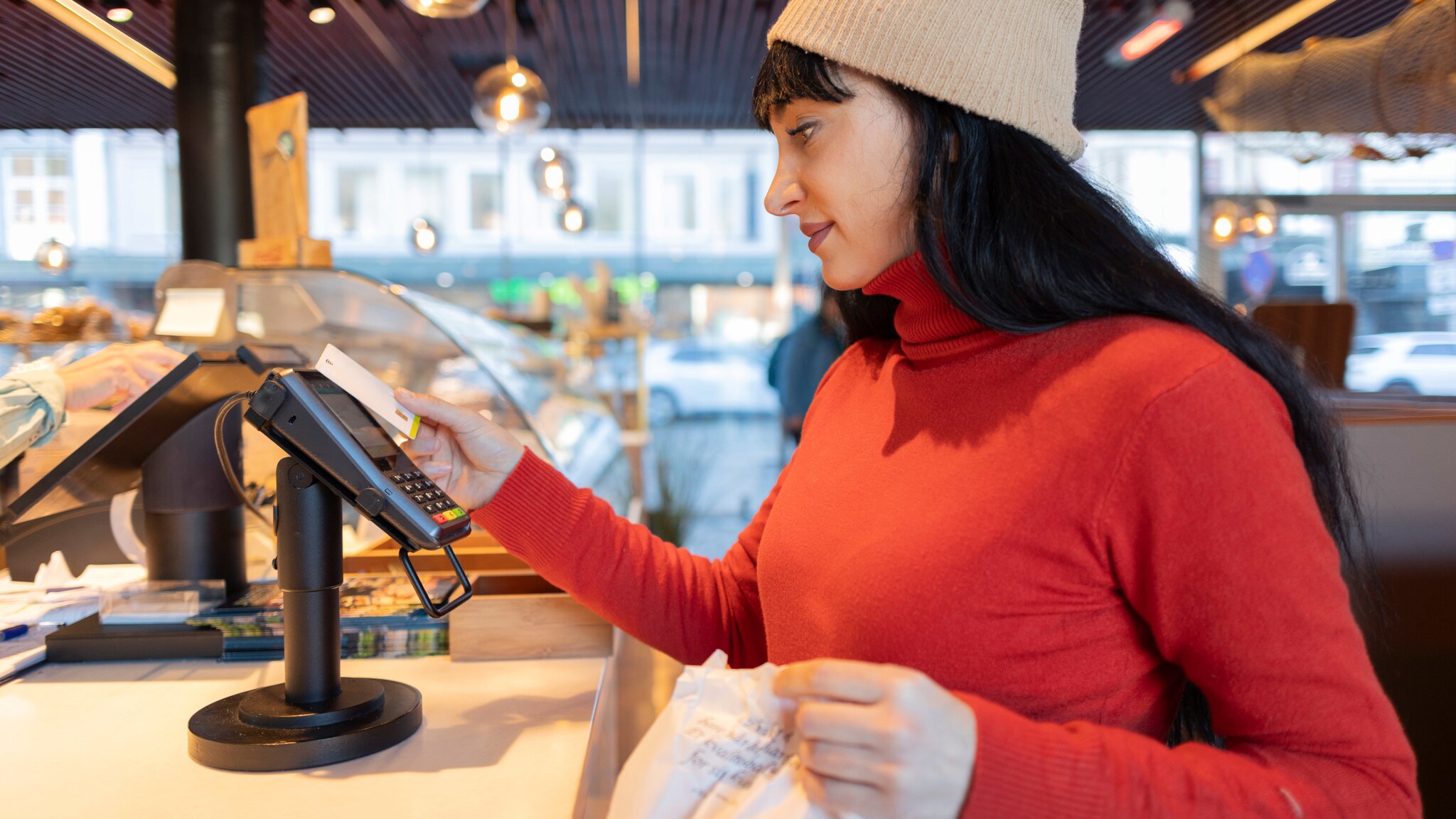 Eine Frau mit Mütze bezahlt mit Kreditkarte an einem Kartenlesegerät in einer Markthalle.