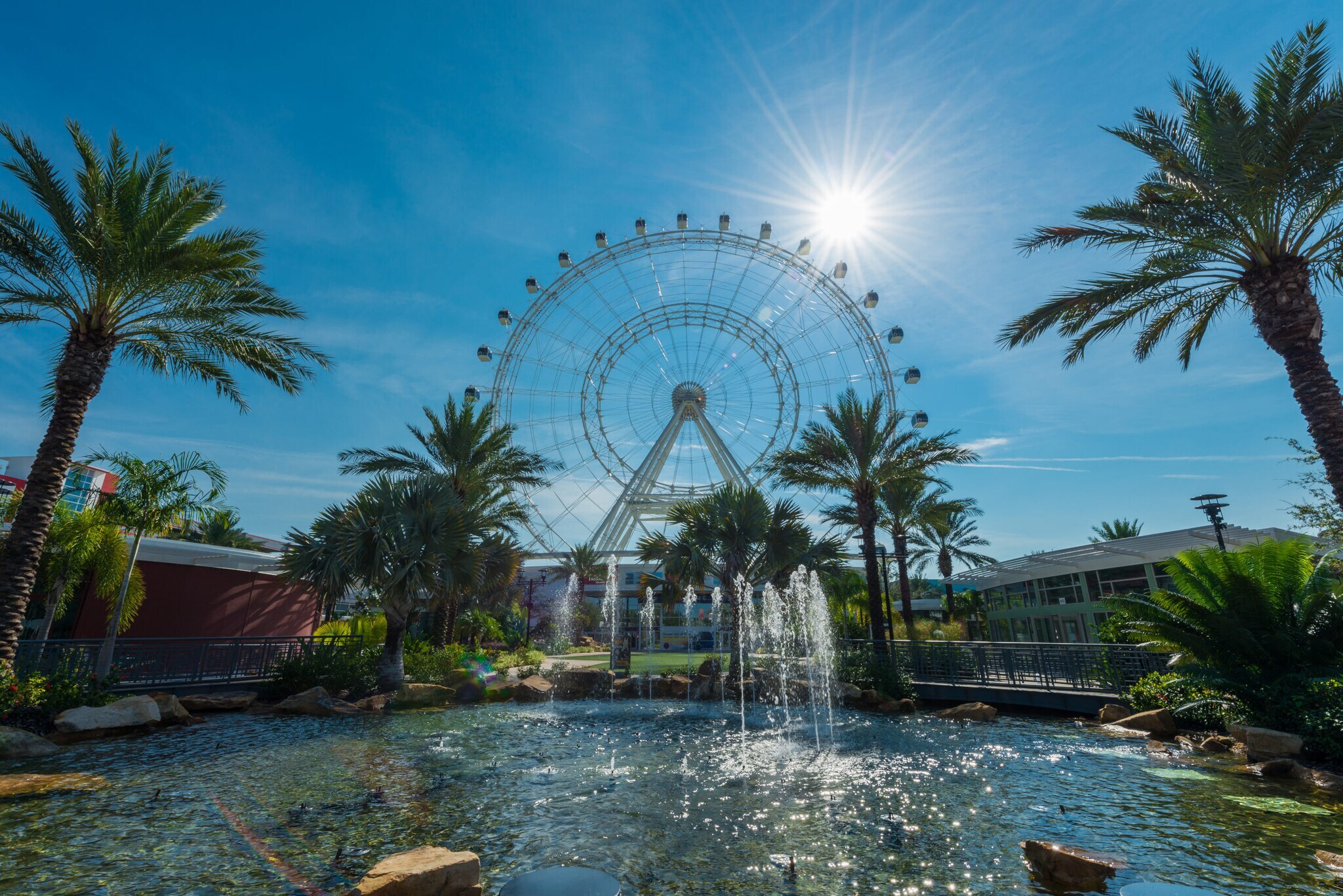 Ein Riesenrad hinter einem Gebäudekomplex mit Gartenanlage samt Teich und Palmen unter blauem Himmel. Ein Riesenrad hinter einem Gebäudekomplex mit Gartenanlage samt Teich und Palmen unter blauem Himmel.
